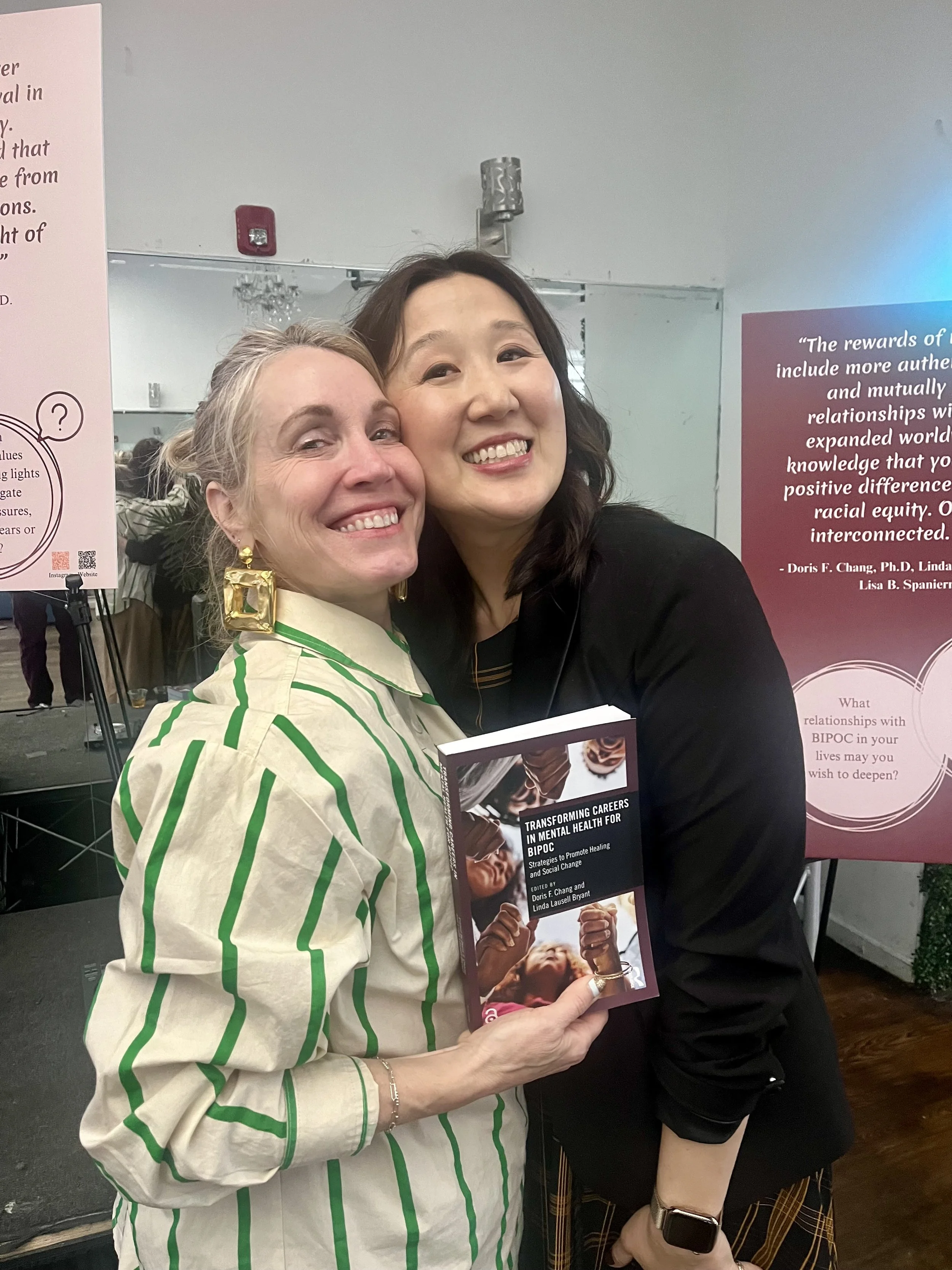 Two women smiling and hugging at a book event, one holding a book titled 'Transforming Careers in Mental Health for BIPOC'.