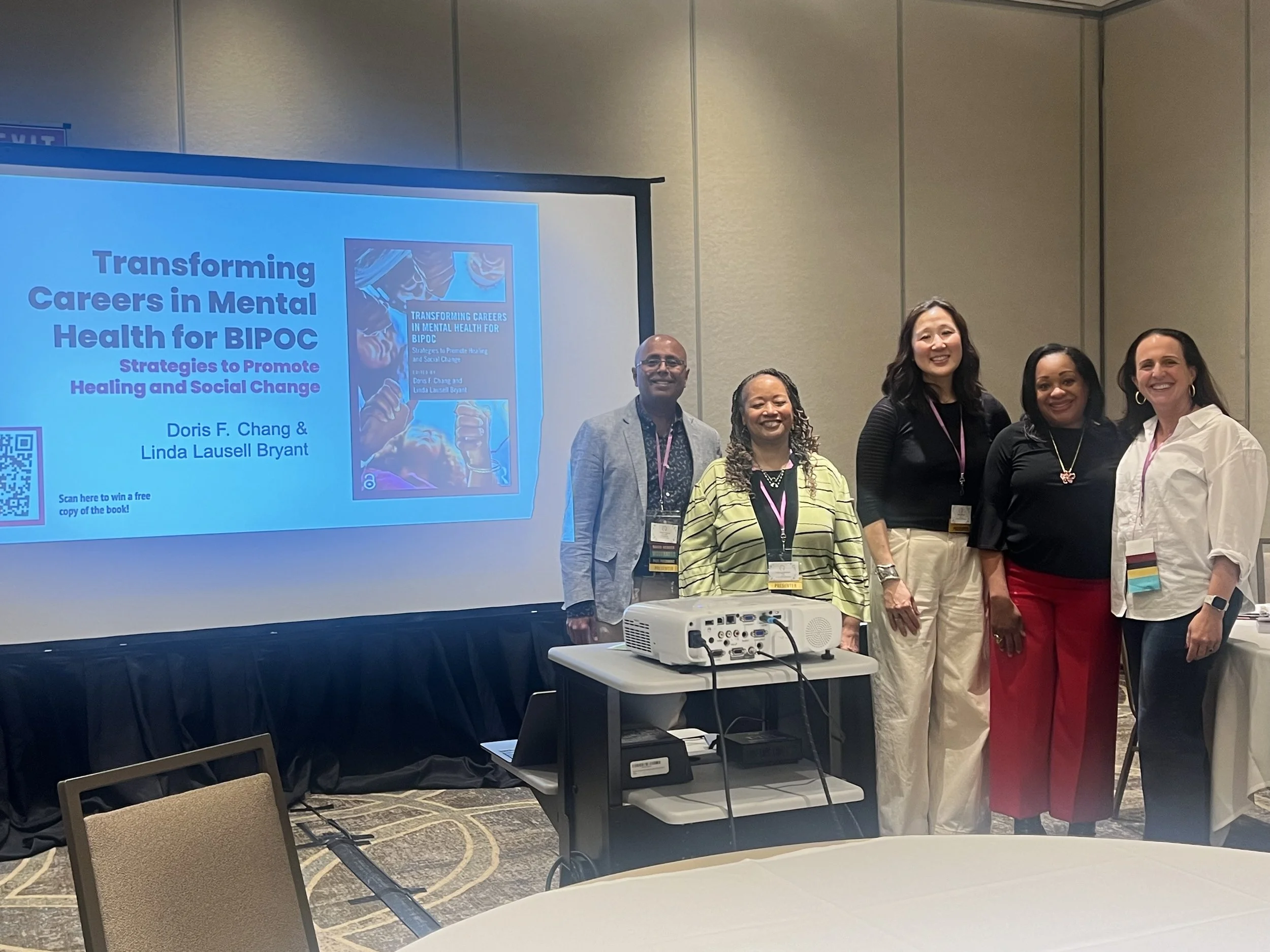 Group of five diverse speakers at a conference, standing in front of a presentation slide about mental health careers for BIPOC, with a projector on a cart in front of them.