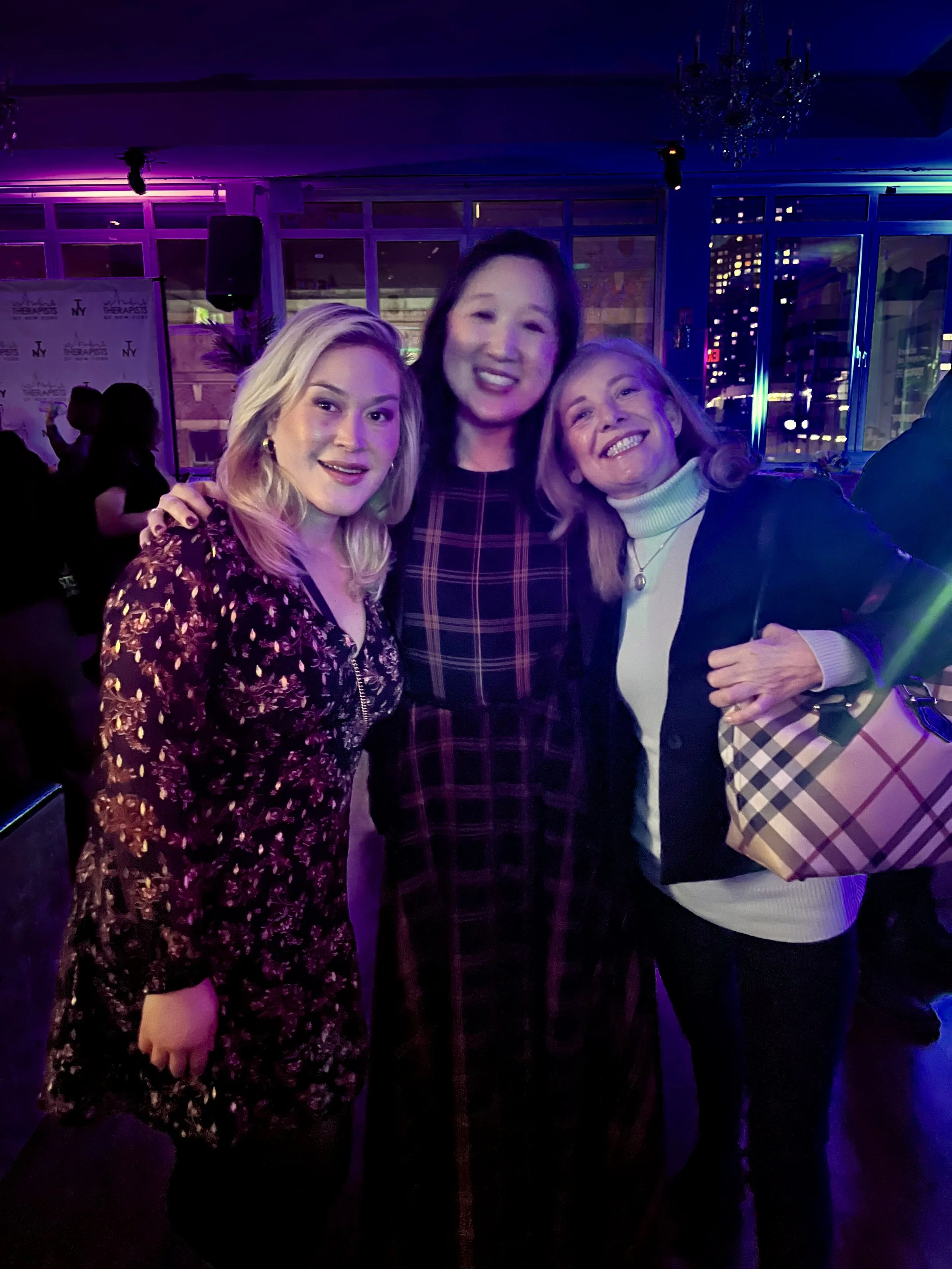 Three women smiling and posing together at an indoor event with city lights visible through windows in the background.