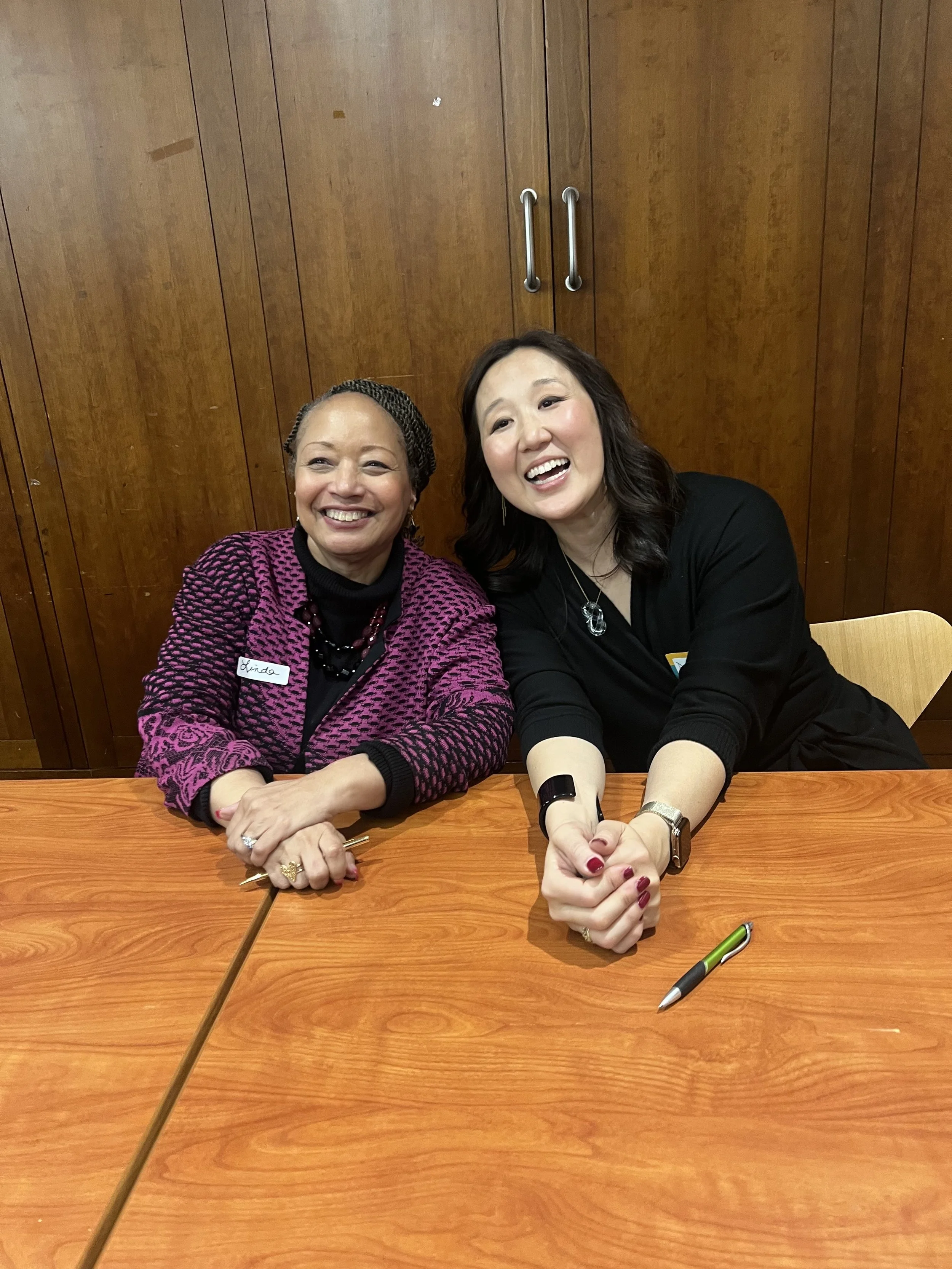 Two women sitting at a wooden table, smiling and leaning towards each other with a wooden cabinet in the background.