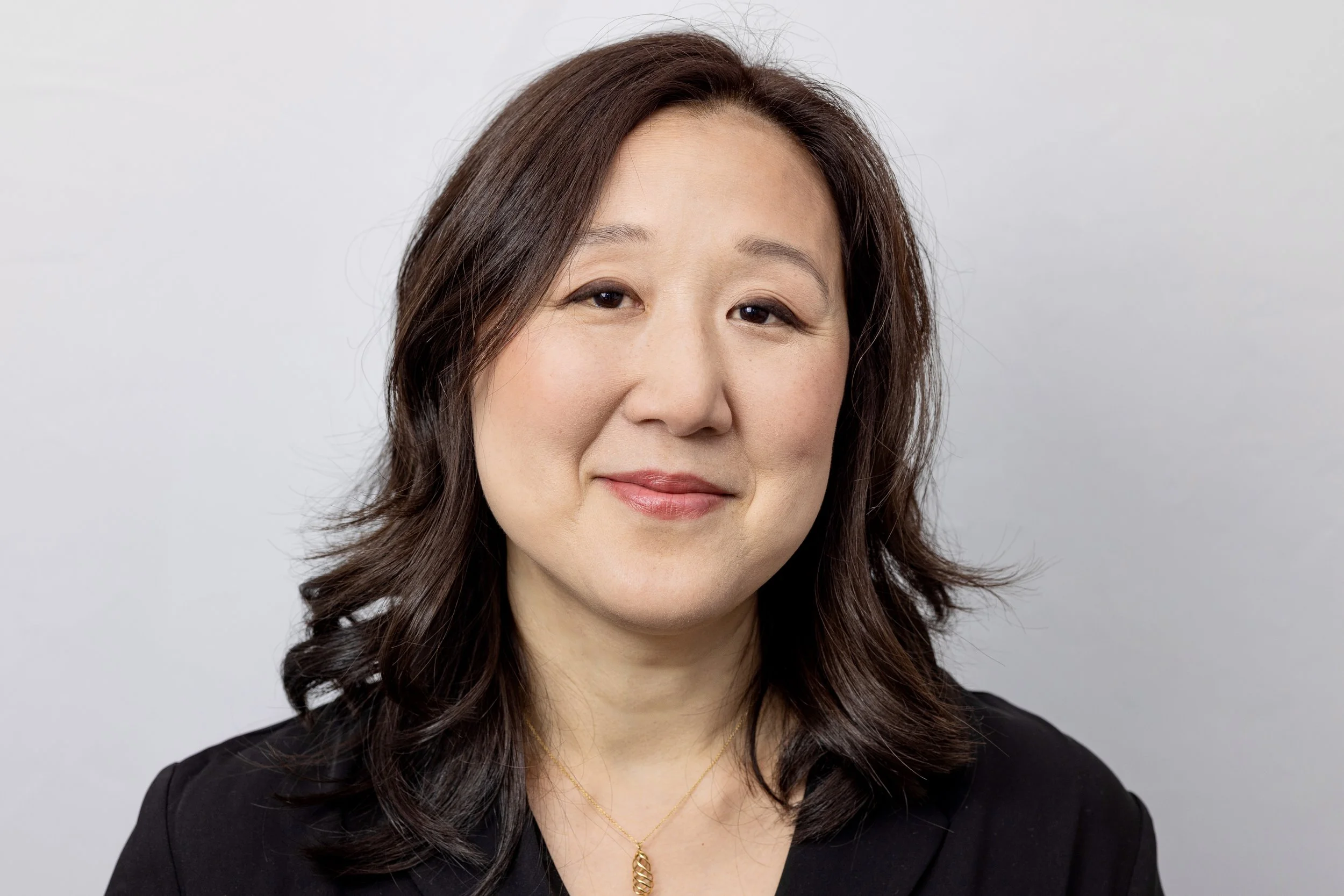 A portrait of a woman with shoulder-length wavy dark hair, wearing a black top and a gold necklace, smiling softly against a plain light-colored background.