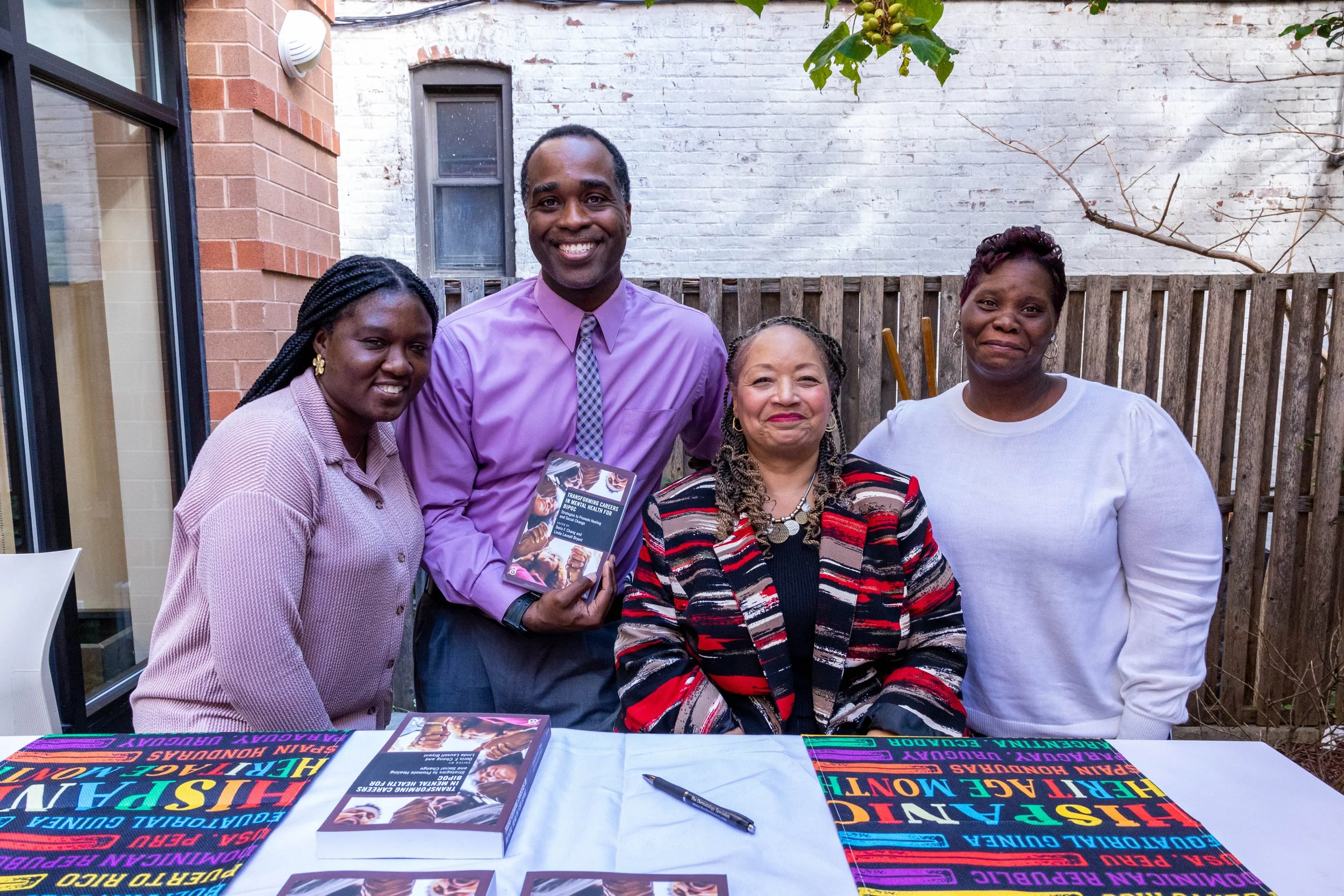Five people standing behind a table outdoors, smiling for the camera. The table has brochures and a colorful cloth with text related to human rights and history.
