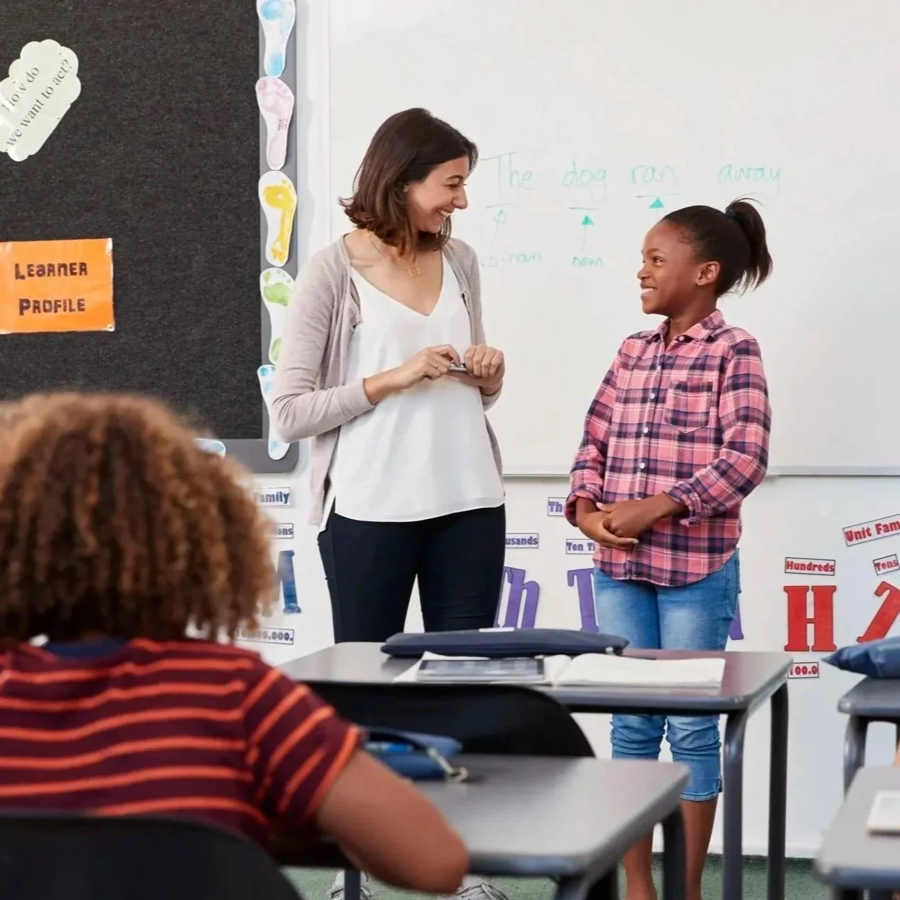 A light-skinned brunette female teacher smiling at a young Black female student at the front of a classroom. The student is standing by a whiteboard, and other students are sitting at desks.