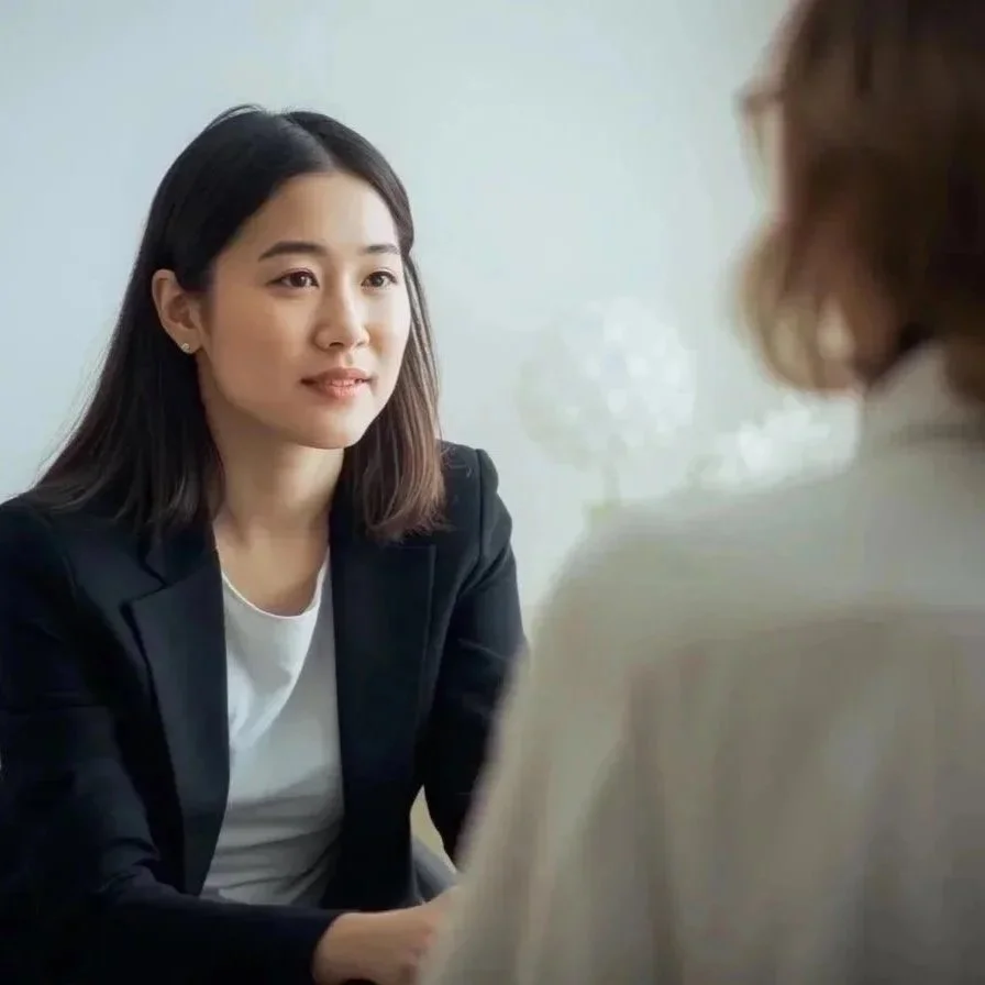 A young Asian woman in a black blazer and white top is having a psychotherapy session with a blond woman whose back is to the camera.