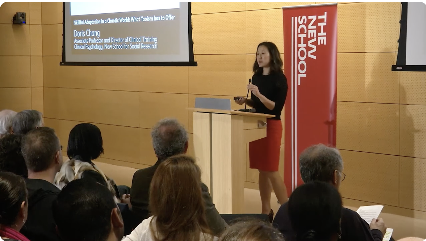 A woman giving a presentation at The New School, standing behind a podium, with an audience seated in front and a large screen displaying her title and credentials.