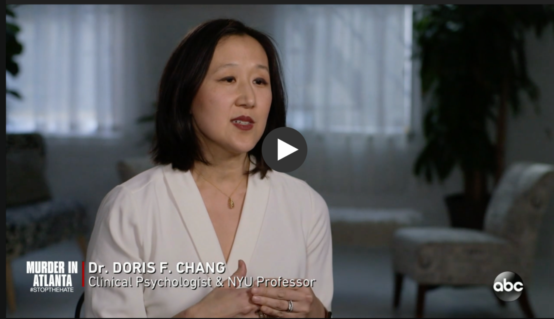A woman with shoulder-length dark hair and light skin, wearing a white top and a gold necklace, speaking in an interview. The background shows a room with large windows, curtains, plants, and furniture. Subtitles identify her as Dr. Doris F. Chang, a clinical psychologist and NYU professor, with a title about murder in Atlanta.