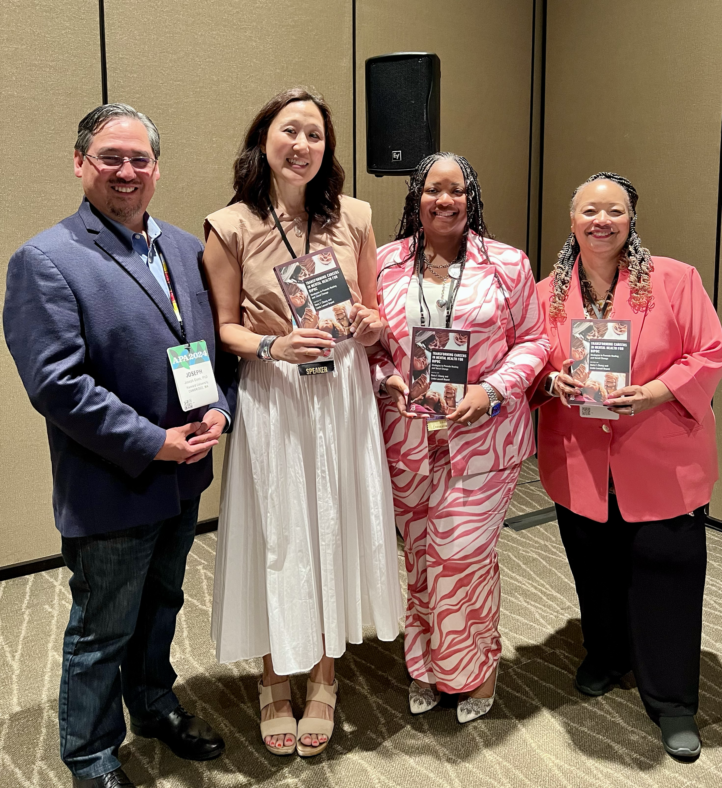 Four people standing together at a conference, holding books and smiling, with a speaker on the wall behind them.