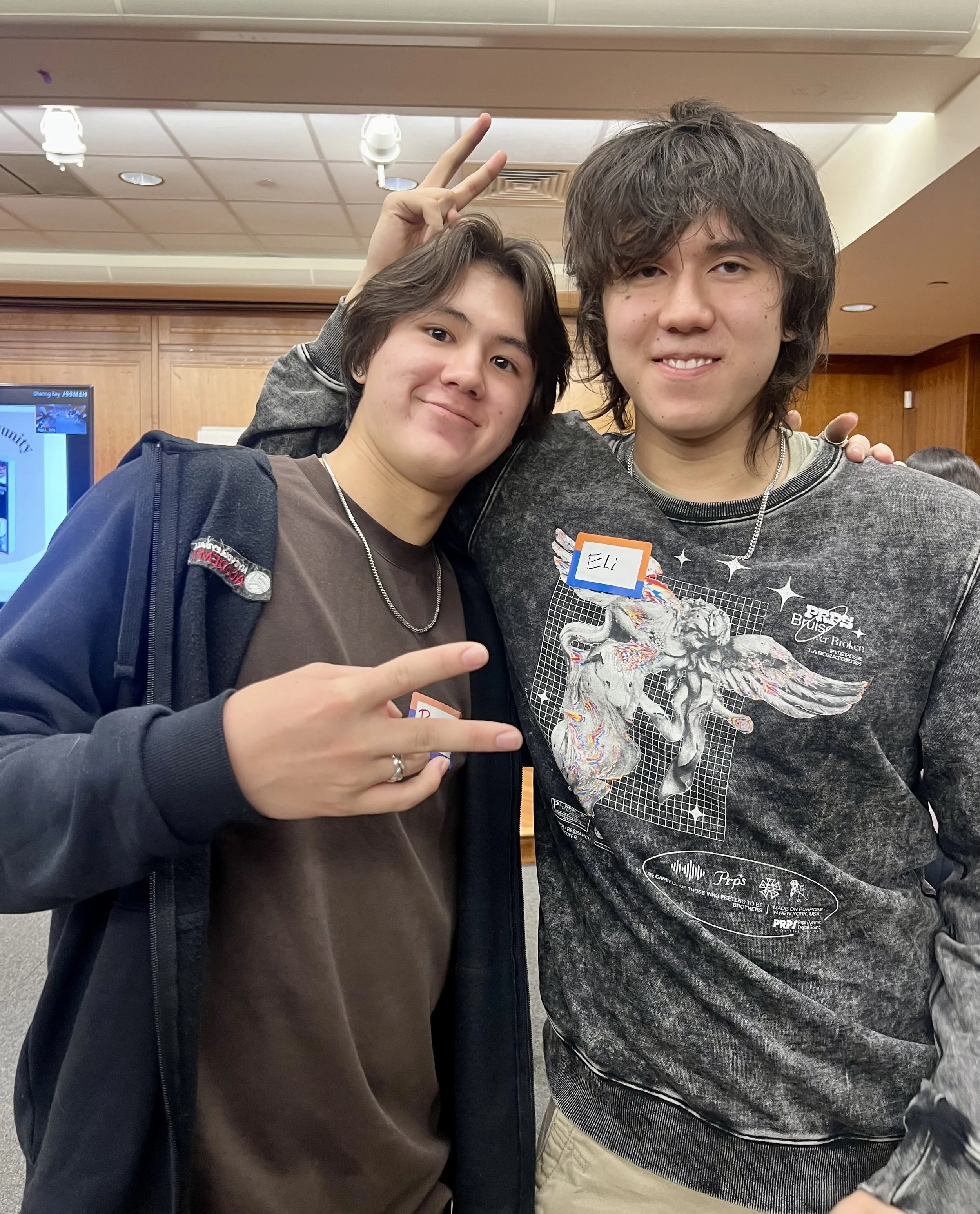 Two young men standing close together in a room, smiling, with one of them making peace signs. The man on the left has dark hair, a brown shirt, and a dark jacket. The man on the right has curly hair, a graphic sweatshirt, and a name tag that says 'E