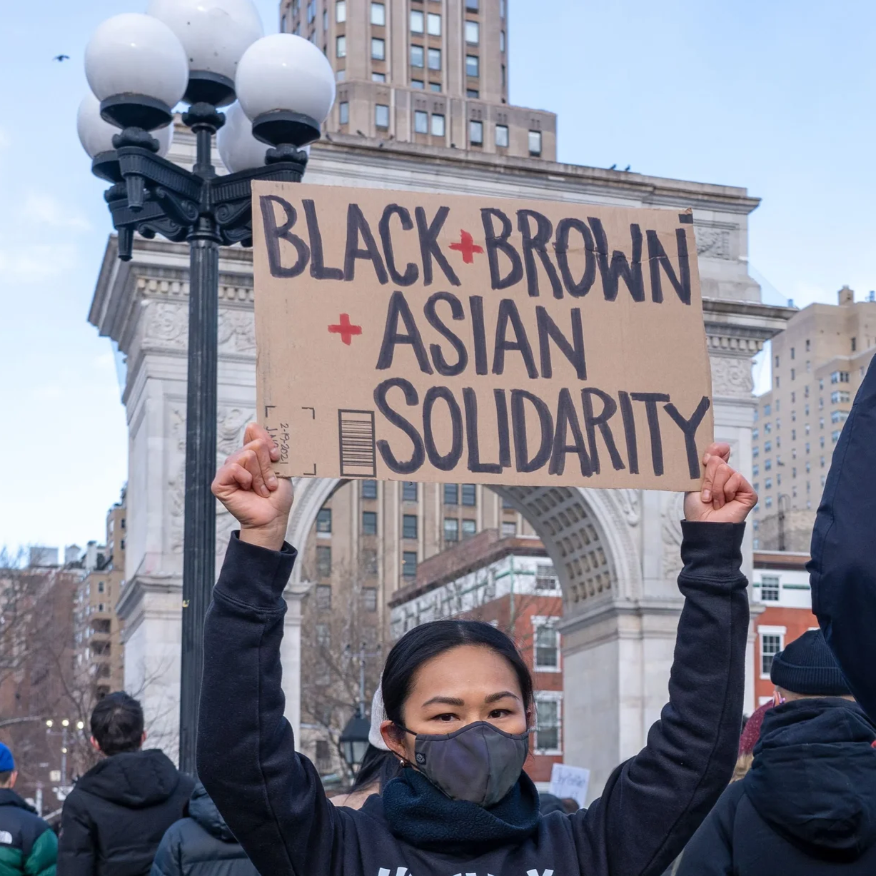 A young woman wearing a face mask holds a sign that reads 'Black + Brown + Asian Solidarity' during a protest in an urban area with tall buildings and a decorative arch in the background.