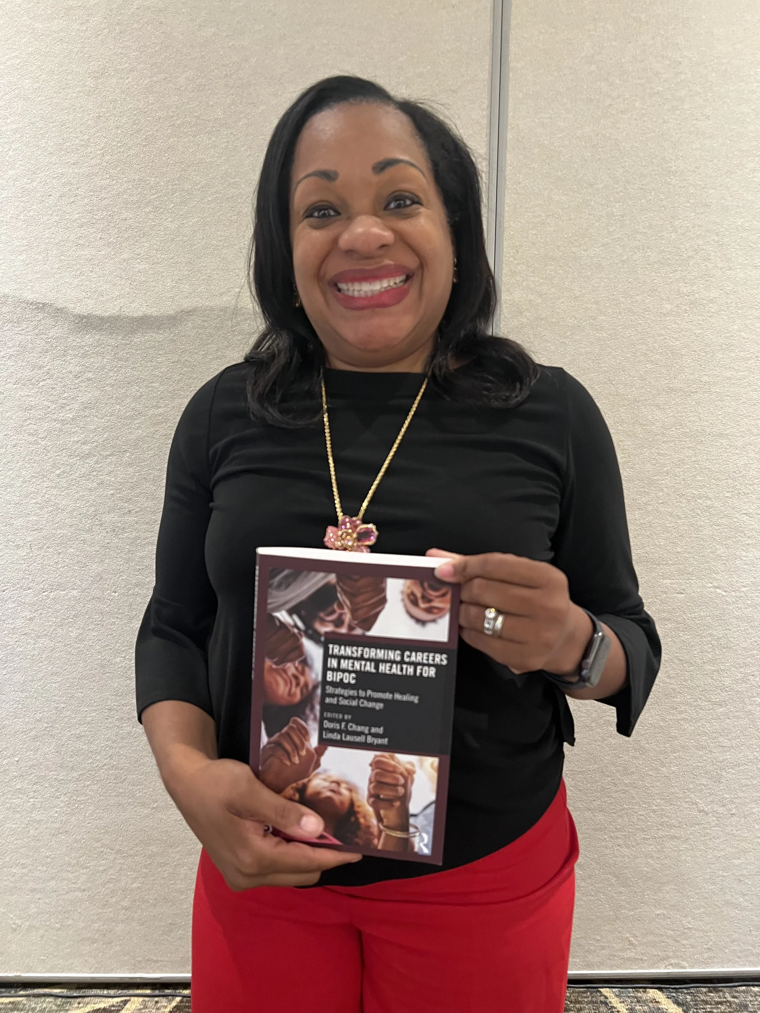 A smiling woman with dark hair, wearing a black top and red pants, is holding a book titled 'Transforming Careers in Mental Health for BIPOC' in front of a neutral wall.