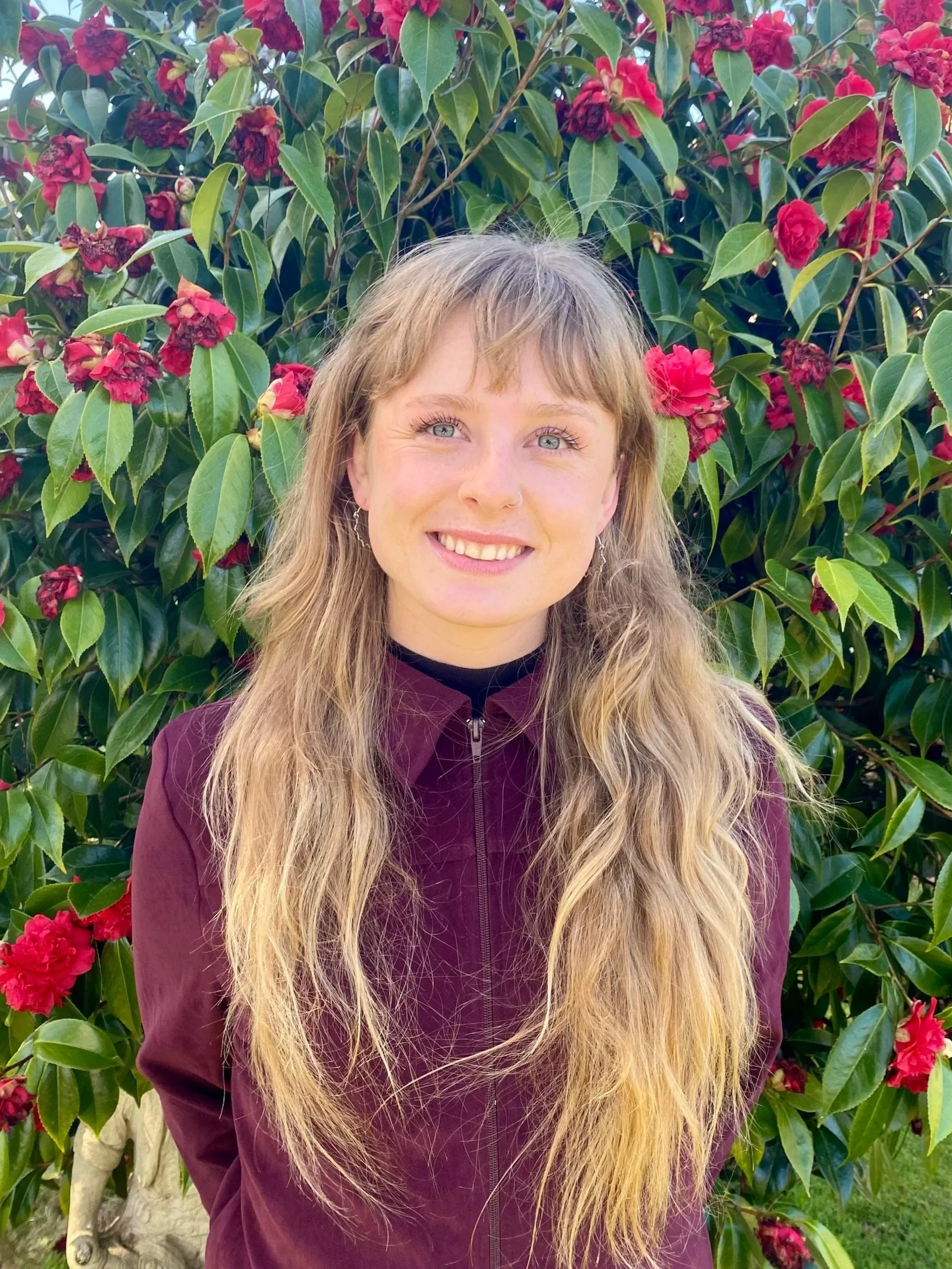 A young woman with long blonde hair and a nose piercing smiling in front of a bush with dark green leaves and pink flowers.