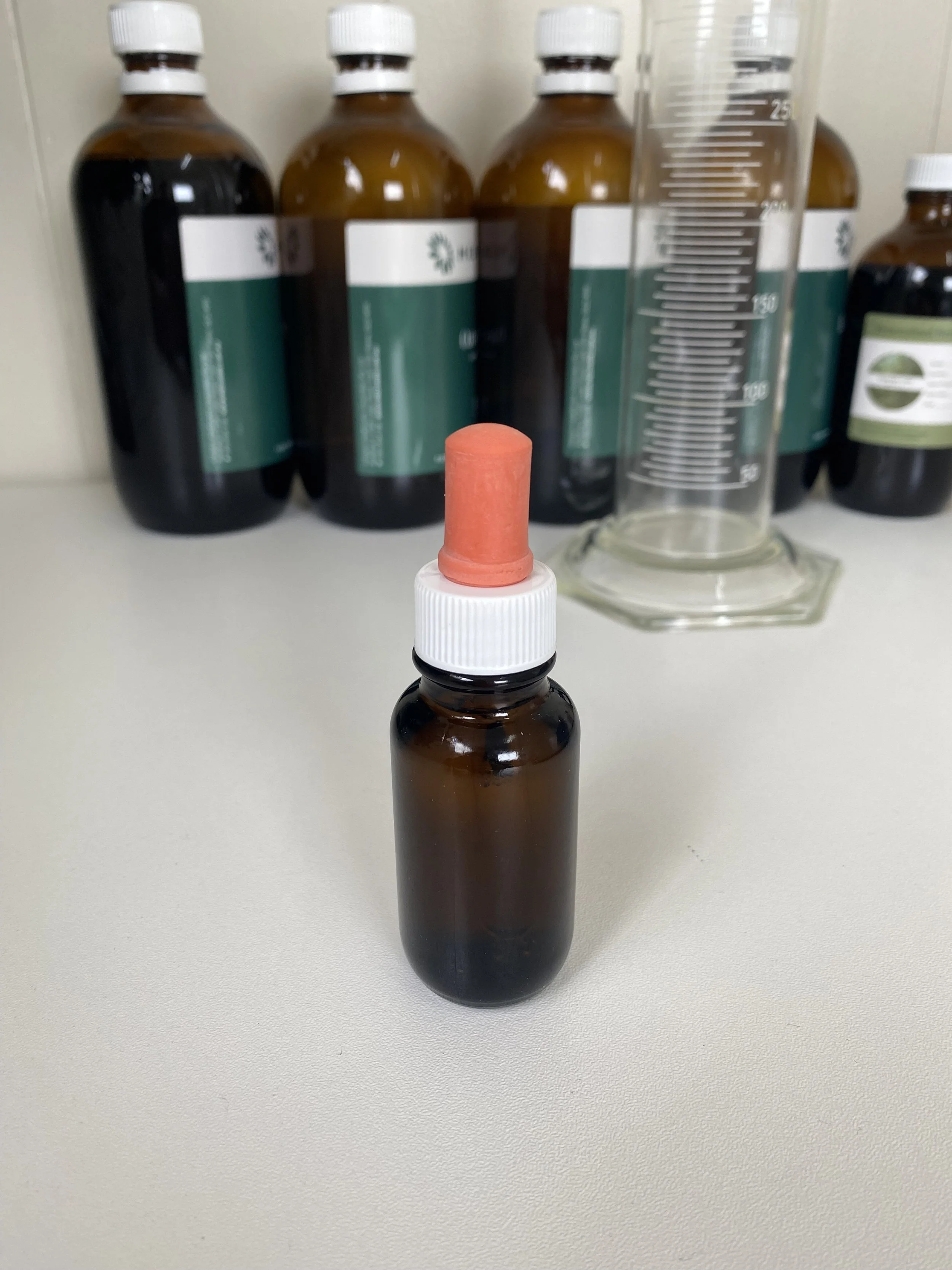 A brown glass medicine bottle with a white screw cap and a red rubber dropper cap, placed on a white surface with larger brown bottles and a graduated cylinder in the background.