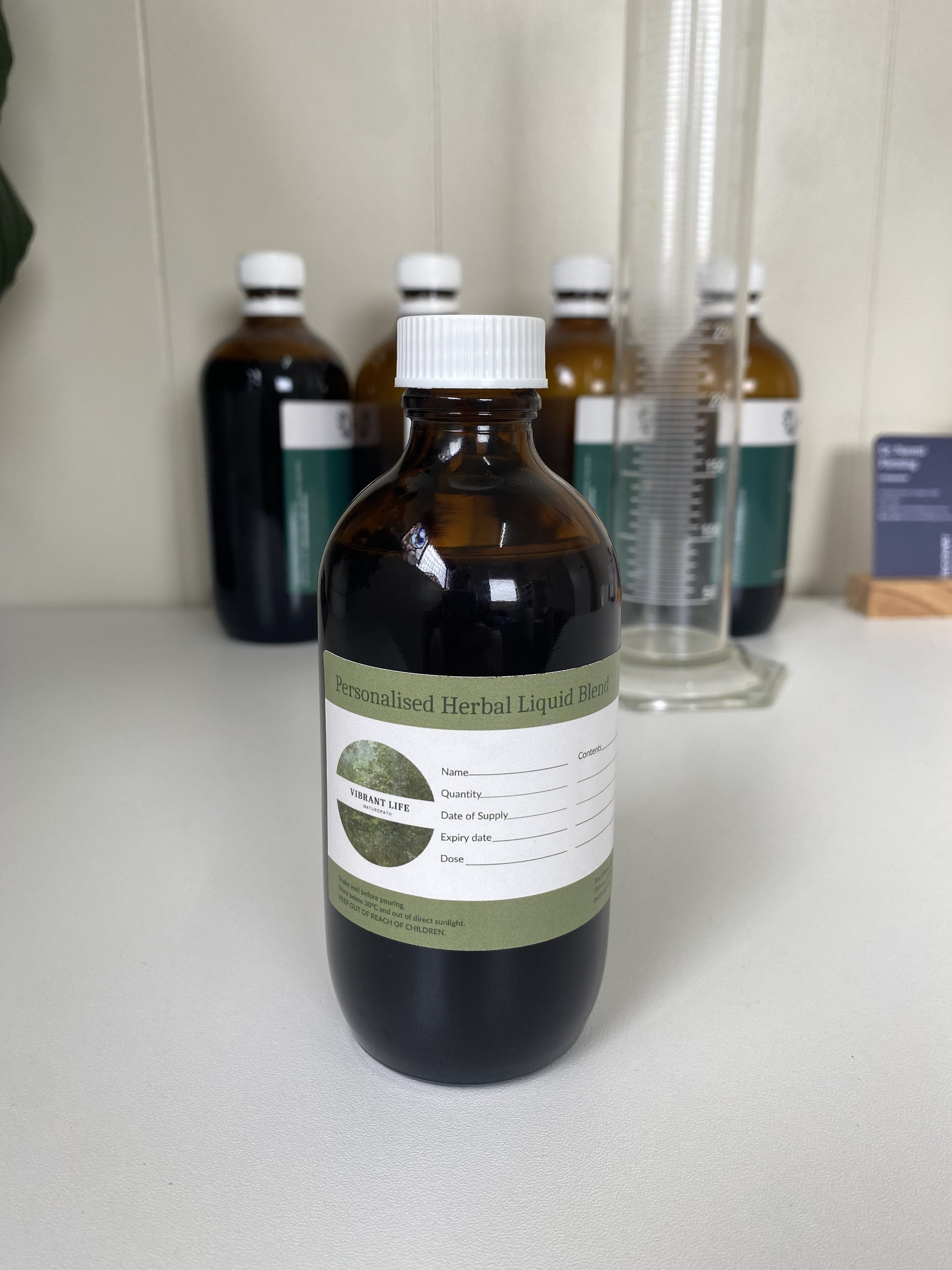 A brown bottle labeled 'Personalized Herbal Liquid Blend' with fields for name, content, quantity, date of supply, expiry date, and dose on a white surface, with other similar bottles and measuring equipment in the background.