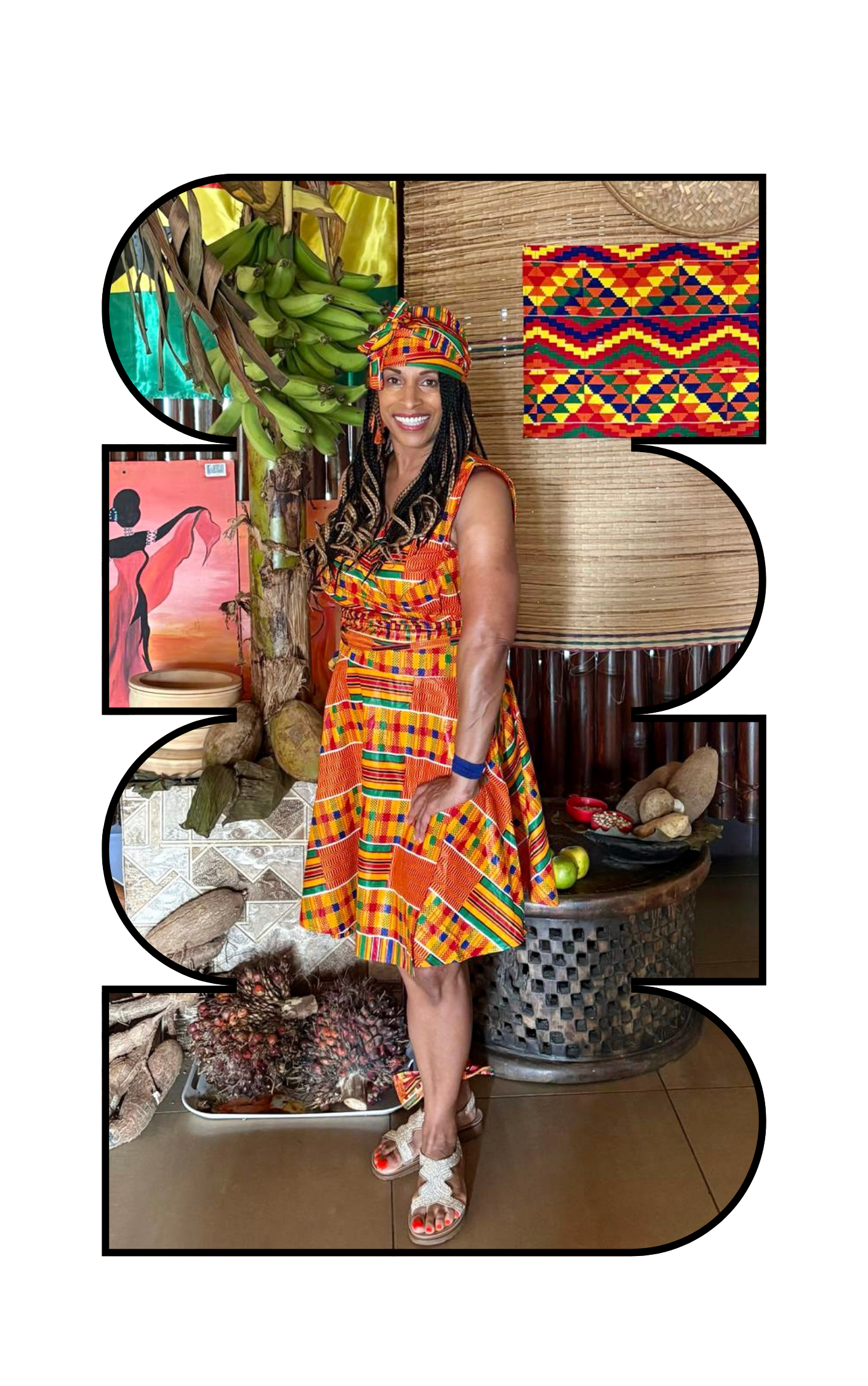 Woman smiling in colorful traditional African dress and headwrap, standing indoors near banana plants and various artwork and decorations.