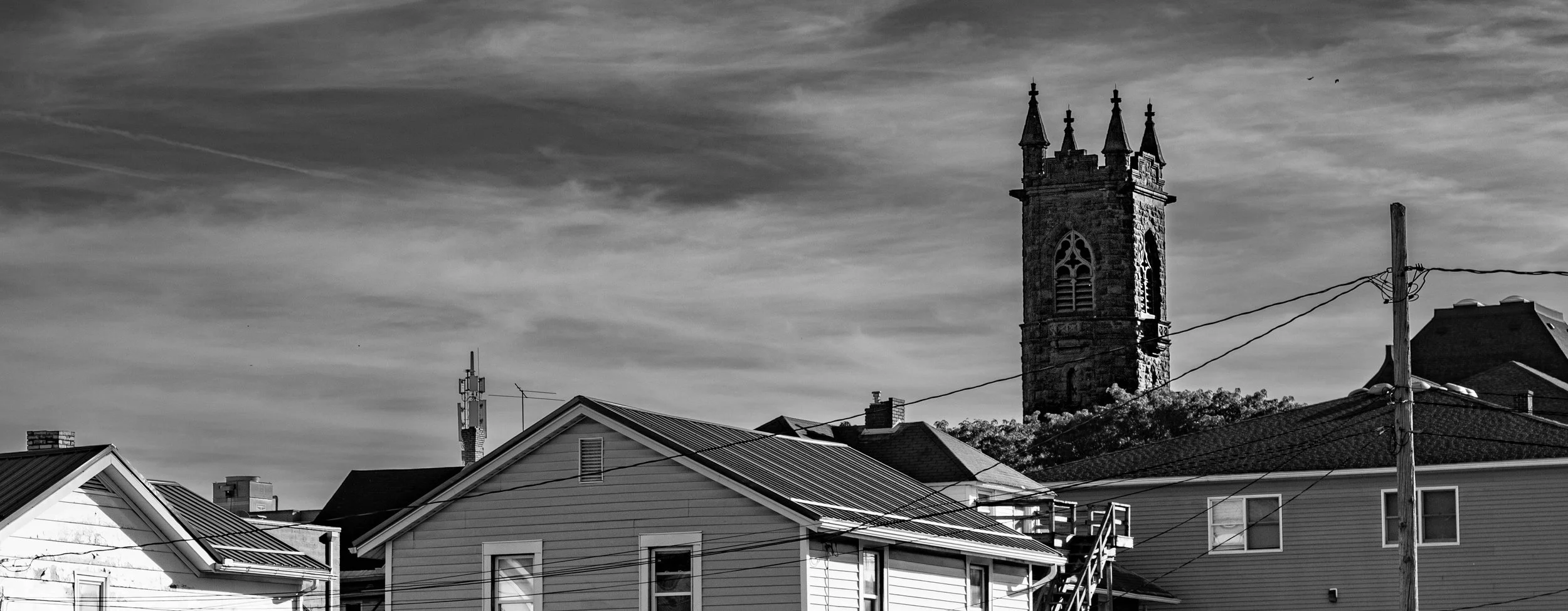 A black and white photograph of a neighborhood with houses, utility poles, and wires in the foreground, and a tall, historic stone clock tower with turrets in the background under a cloudy sky.