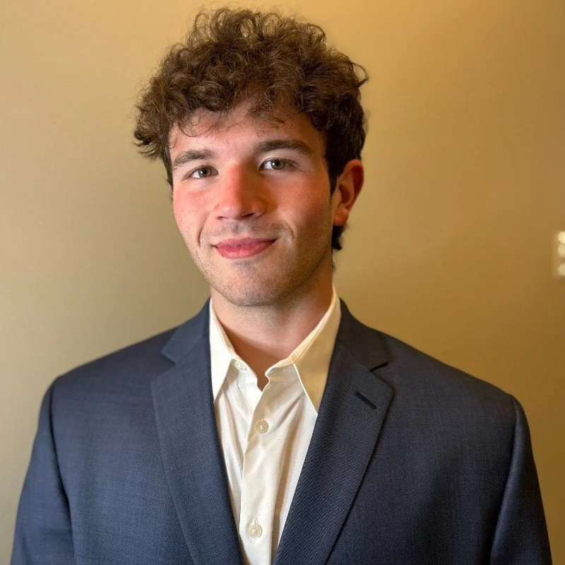 A young man with curly brown hair dressed in a dark suit and a white shirt, standing against a beige wall with a soft smile.