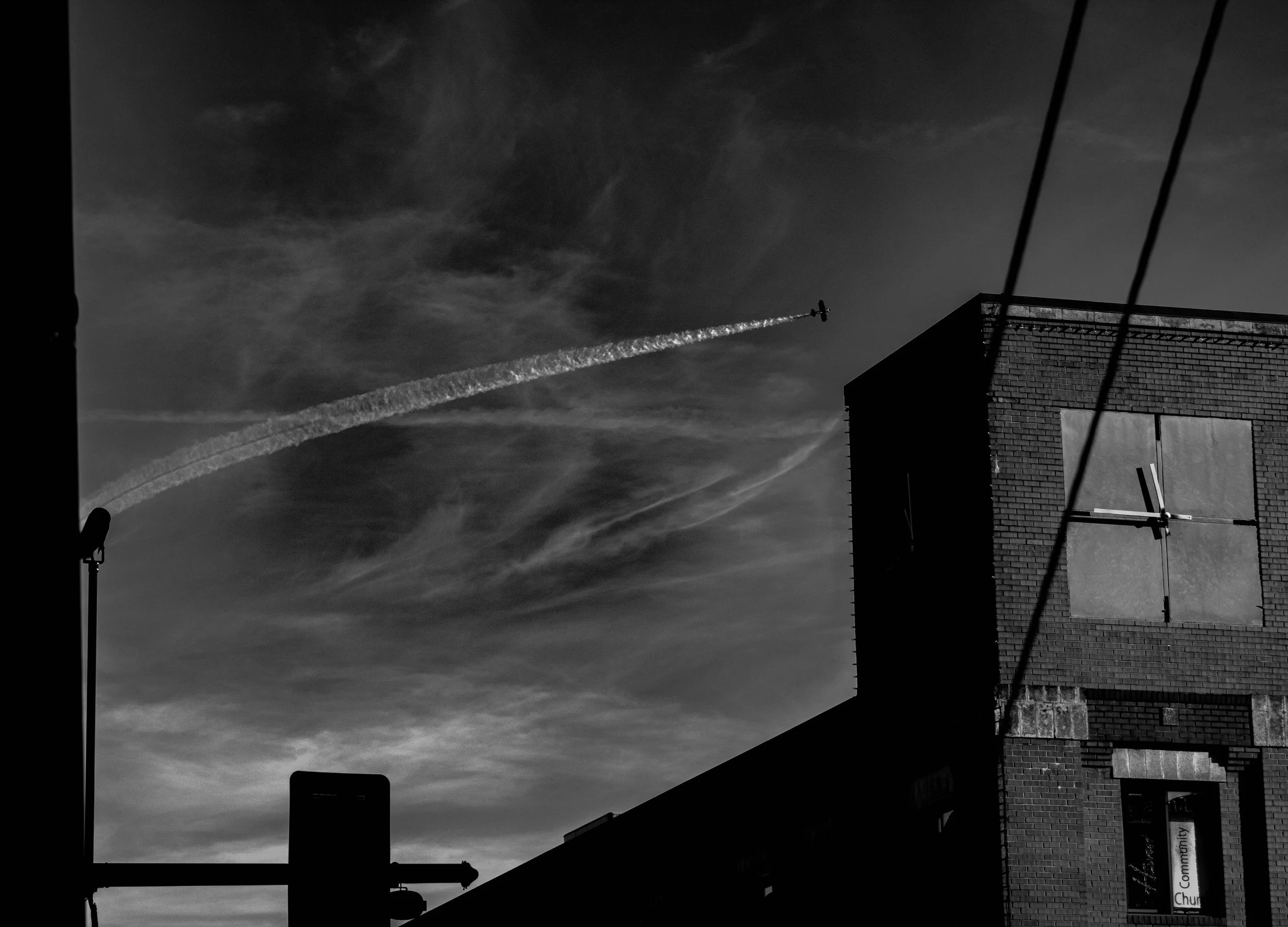 Black and white photo of urban buildings, with a small airplane flying in the sky leaving a contrail, and a streetlamp silhouette in the foreground.