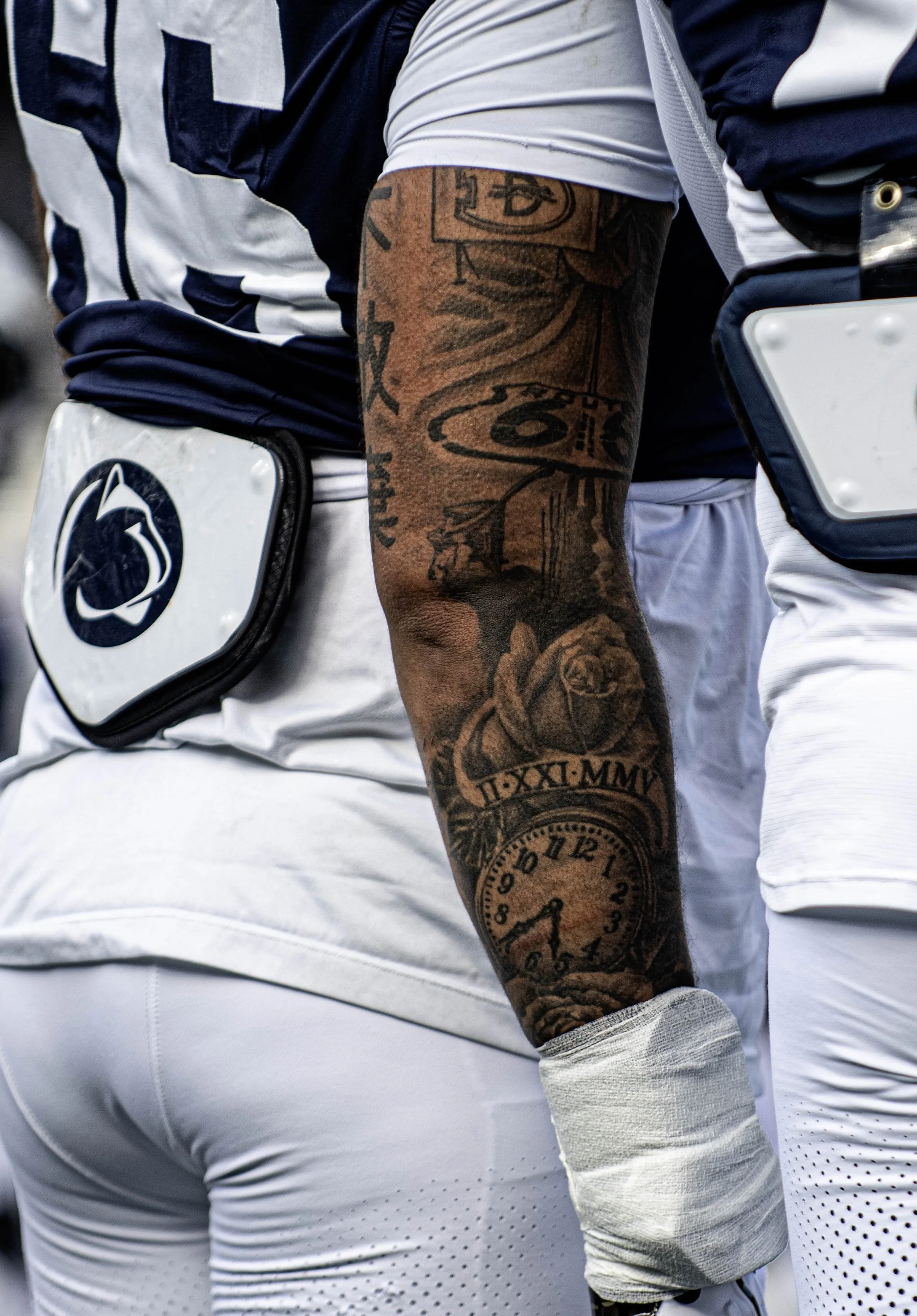 Close-up of a football player wearing a navy and white uniform with a tattooed arm and patch on the back, standing next to another player in similar uniform.