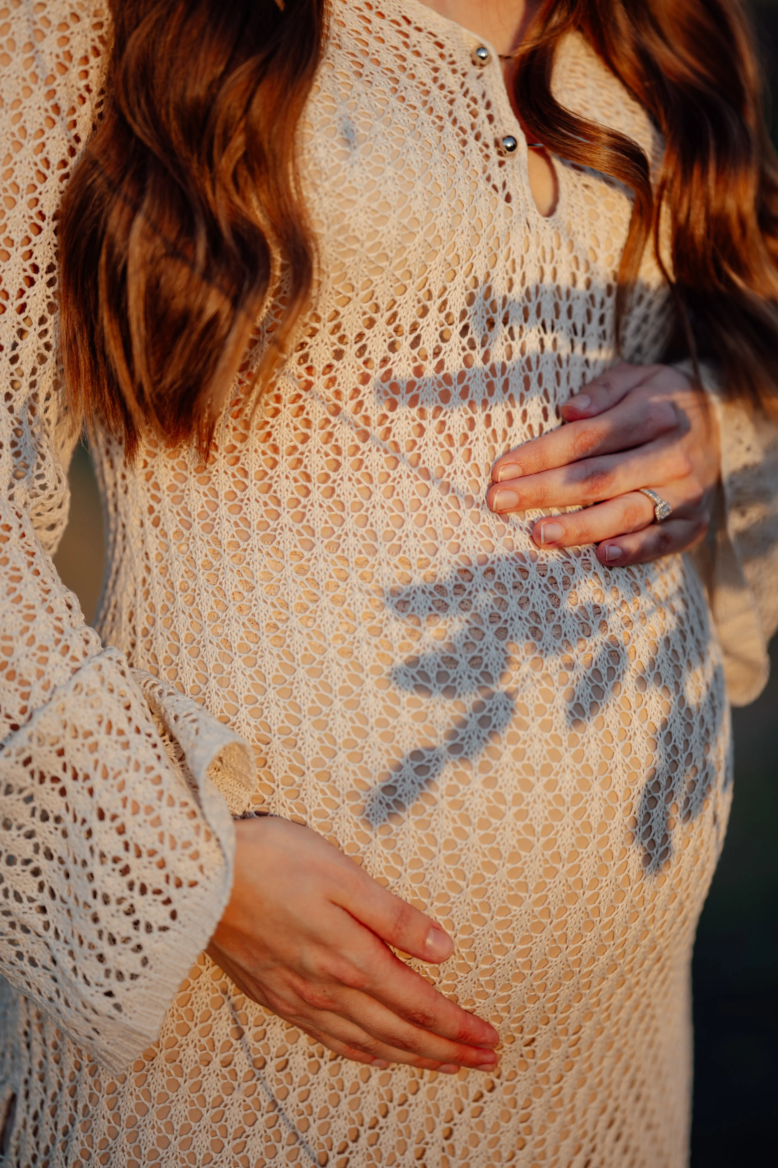 Close-up of a pregnant woman wearing a cream-colored, lace top, gently holding her baby bump with both hands, one hand displaying an engagement ring. The sunlight creates shadows on her belly from her hands and hair.