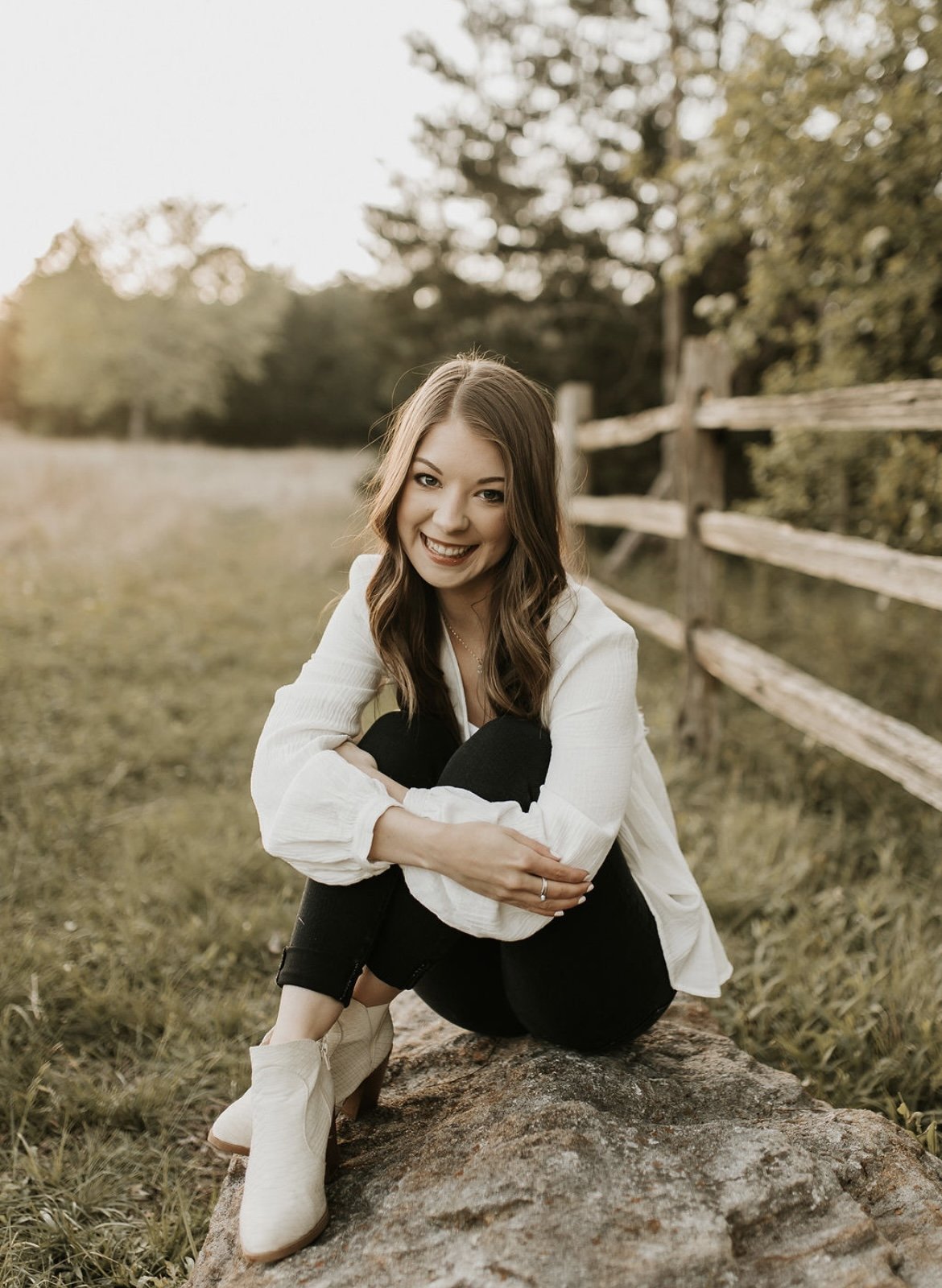 A young woman with brown hair, smiling, sitting on a large rock in a grassy outdoor area during sunset, wearing a white blouse, black pants, and beige boots, with a wooden fence and trees in the background.