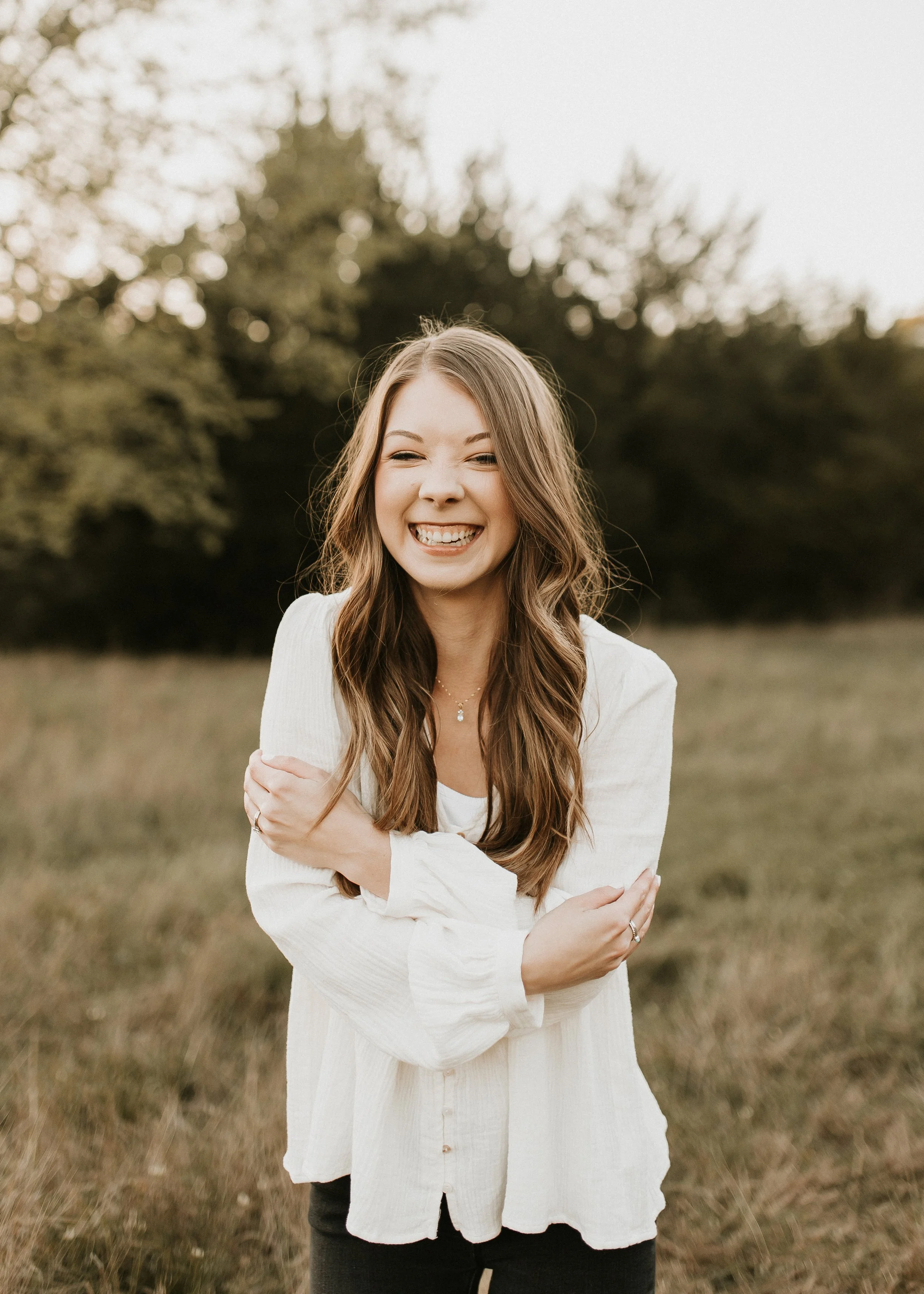 A woman with long brown hair smiling outdoors in a field with trees in the background, wearing a white blouse.
