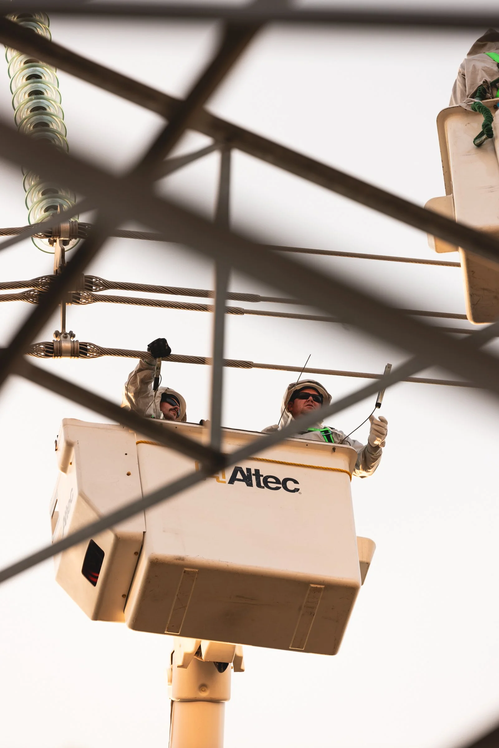 Two utility workers in a bucket lift working on power lines, visible through grated metal fencing.