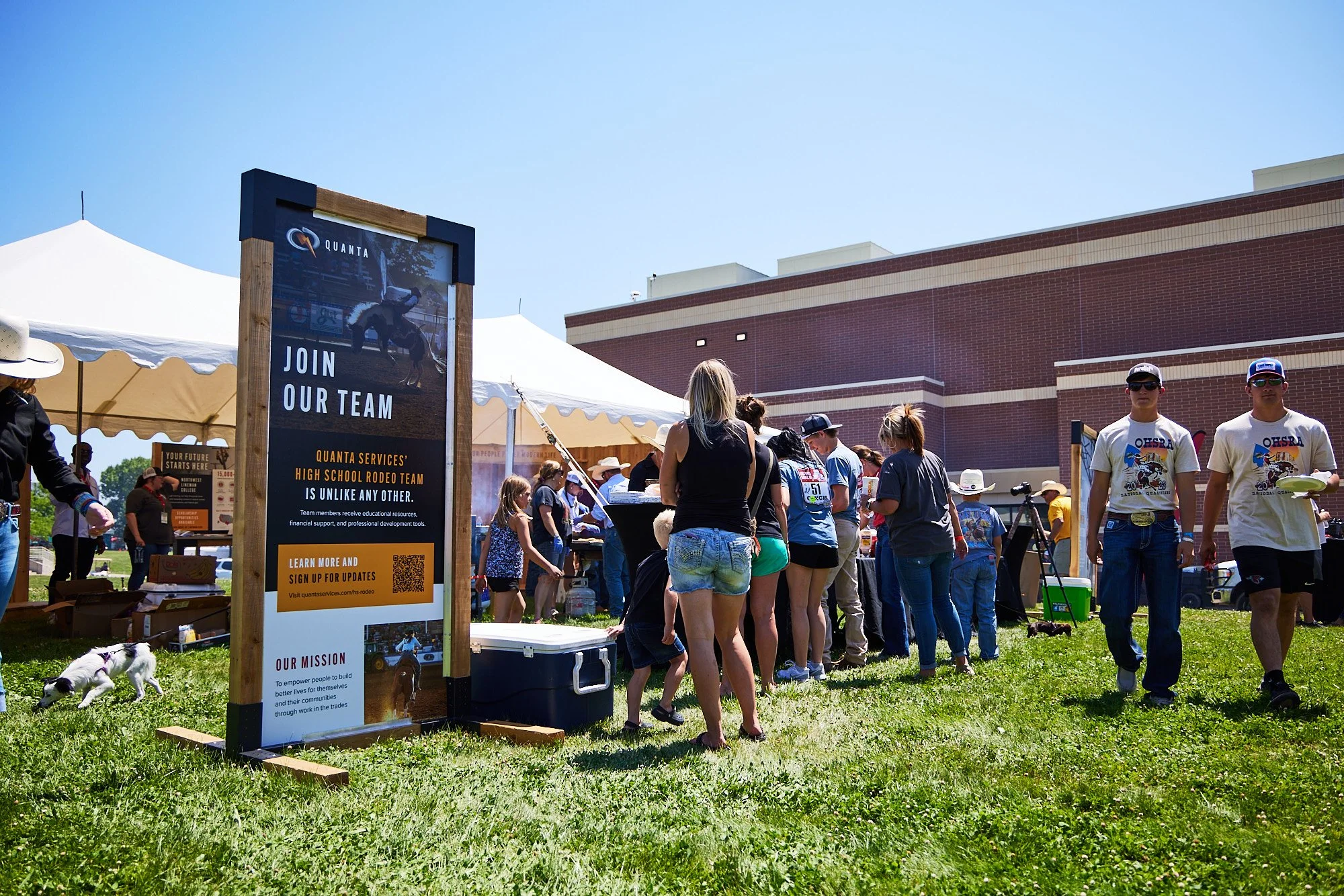 People standing in line at an outdoor event with tents and a large sign promoting a high school rodeo team, on a sunny day.