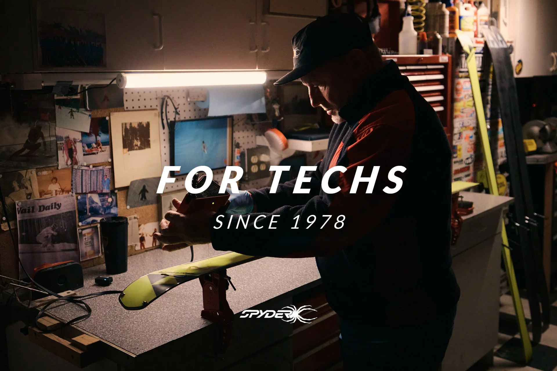 A person working in a cluttered workshop, holding a device, surrounded by tools, photos, and equipment, with the text 'For Techs Since 1978' overlaid.