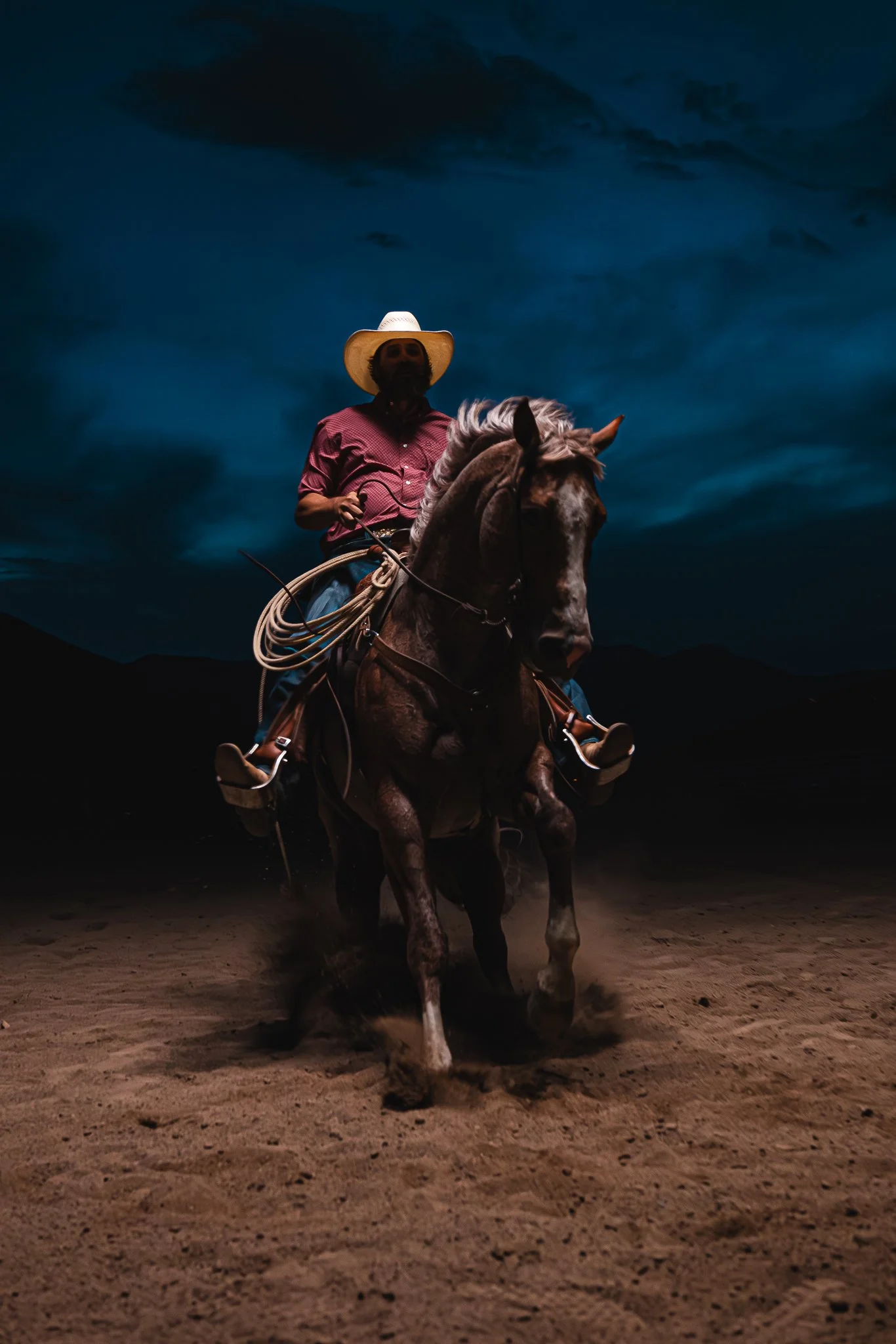 A person riding a horse on a dirt ground during night time, with dark clouds in the sky.