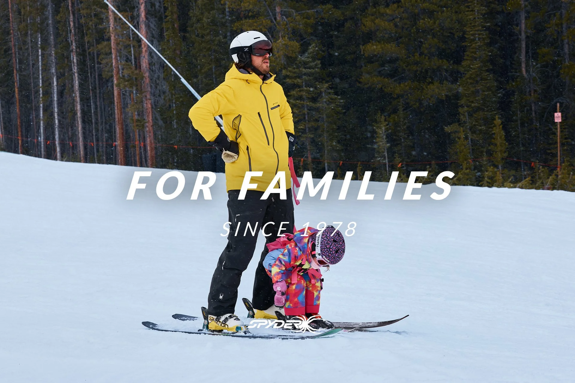 An adult man and young girl skiing on snow with a forested background. The man is wearing a yellow jacket, black pants, a white helmet, and goggles. The girl is dressed in colorful winter clothing and a helmet, sitting on skis.