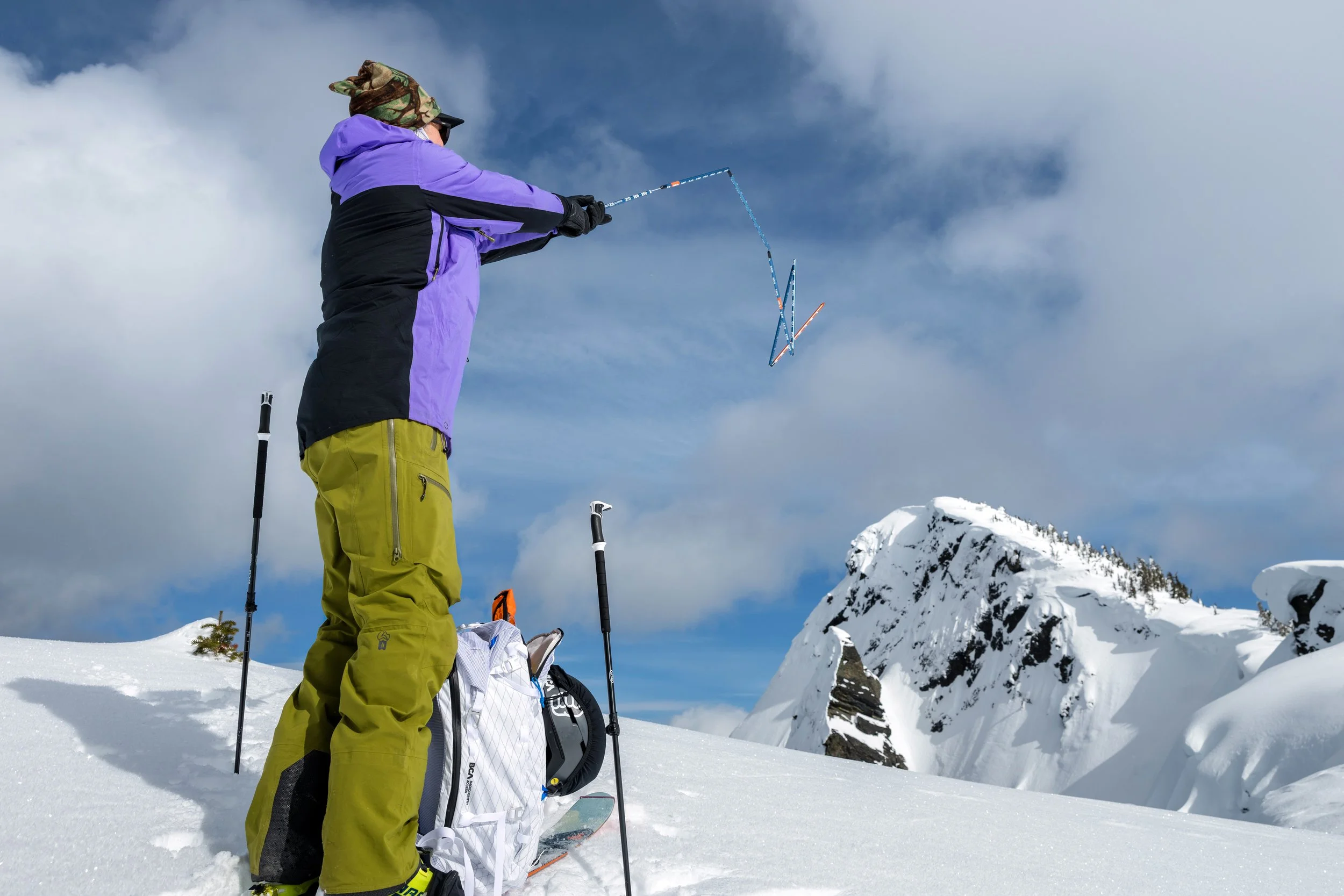 A person in winter outdoor gear casting a snow shape with a stick on a snowy mountain landscape.
