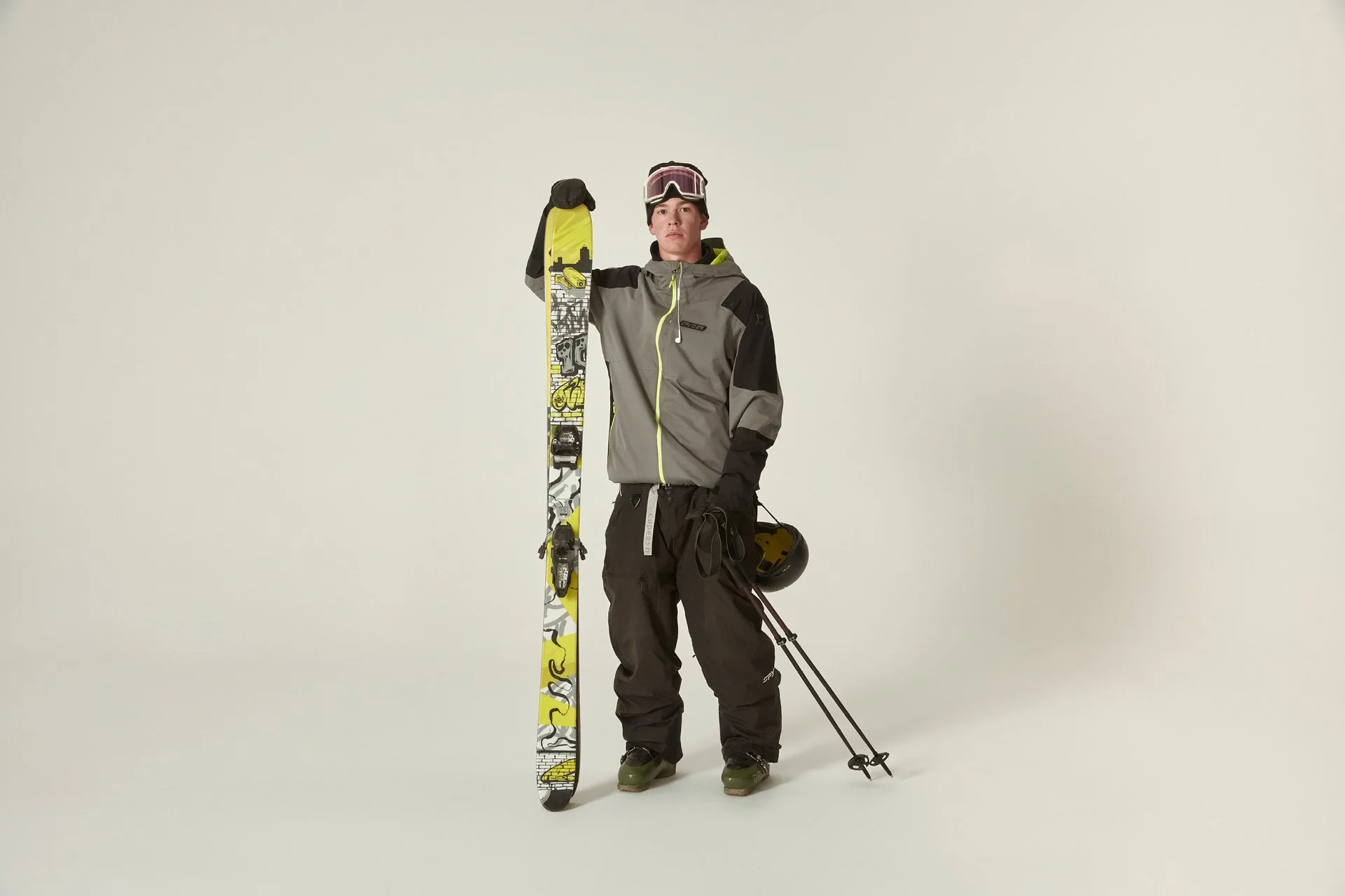 A man dressed in ski gear standing against a plain white background, holding a pair of skis in one hand and ski poles in the other, with a ski helmet and goggles.