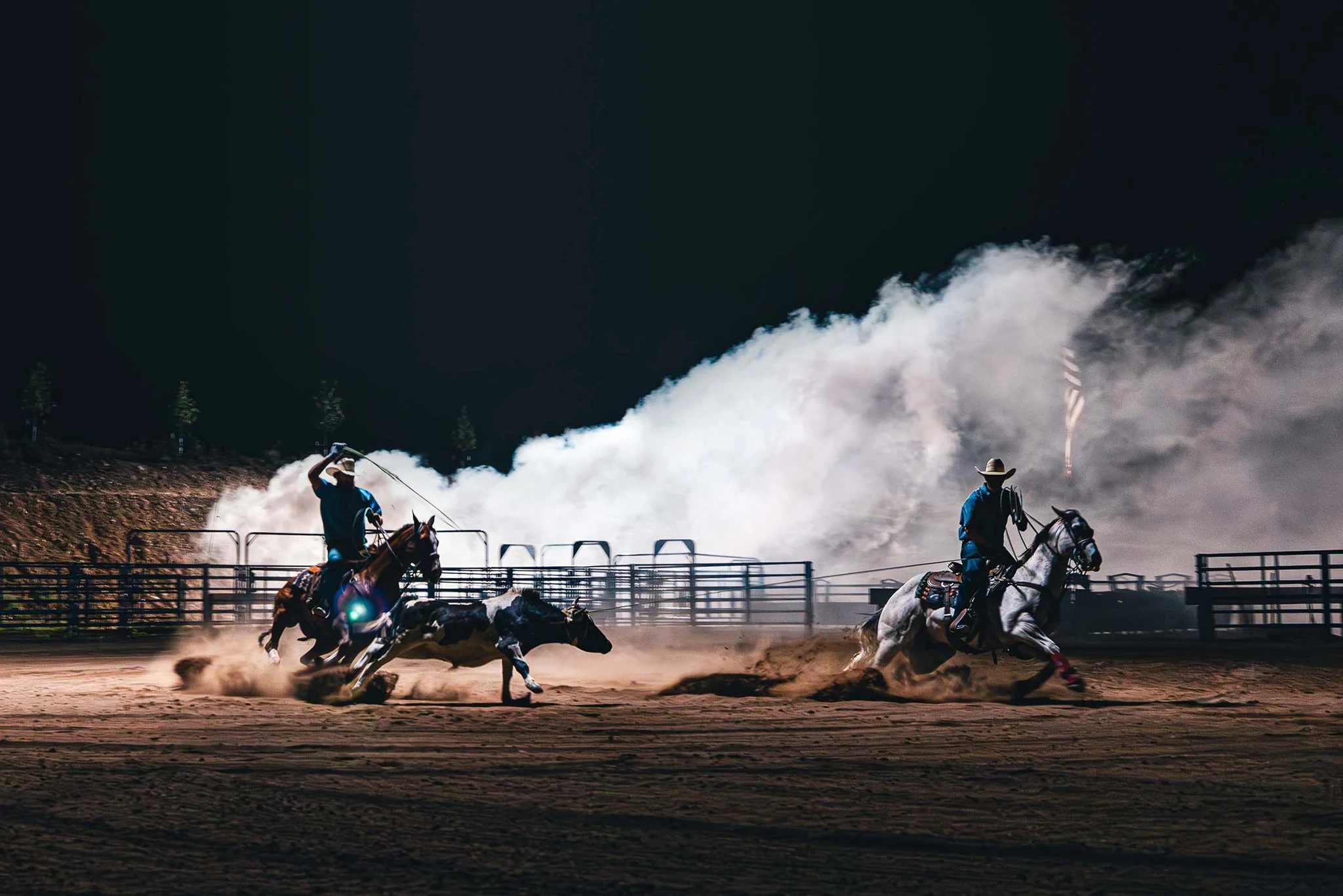 Two cowboys riding horses on dirt arena at night, chasing a calf, with a large cloud of dust behind them.