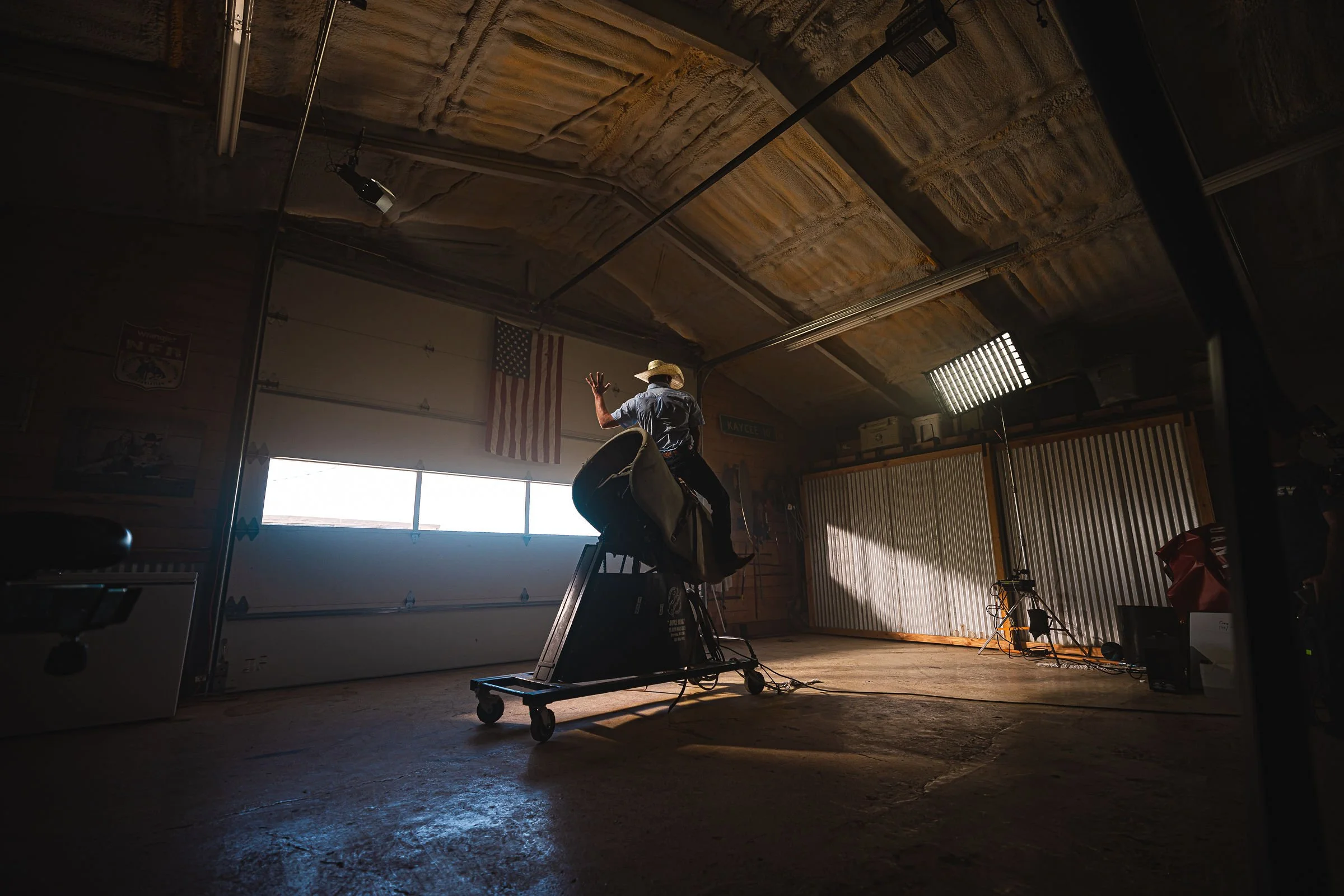 A person wearing a cowboy hat and jeans riding on a treadmill that is set up inside a garage, with American flags and various garage items in the background.