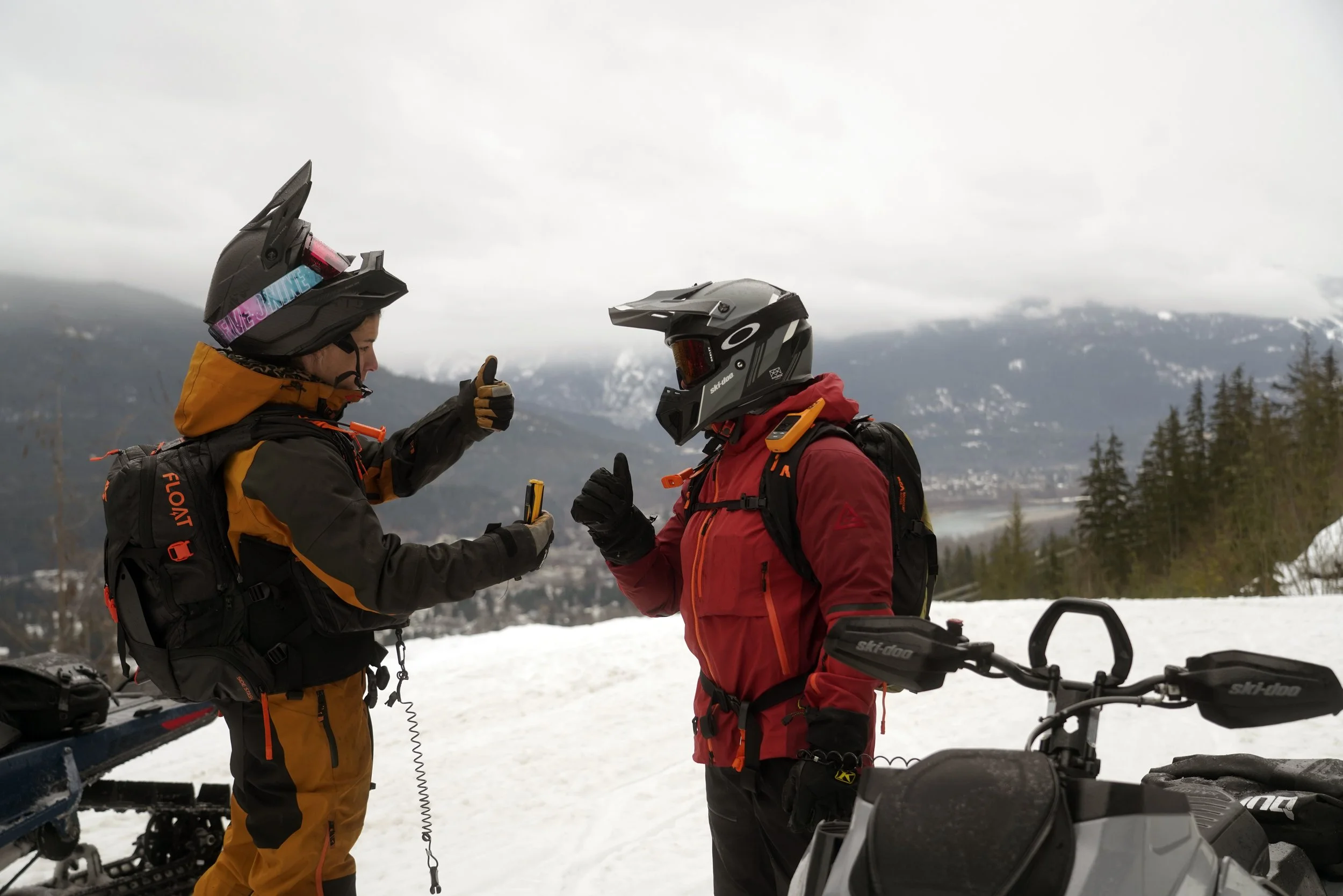 Two people in snow gear and helmets talking outdoors near snowmobiles, with a mountain landscape and cloudy sky in the background.
