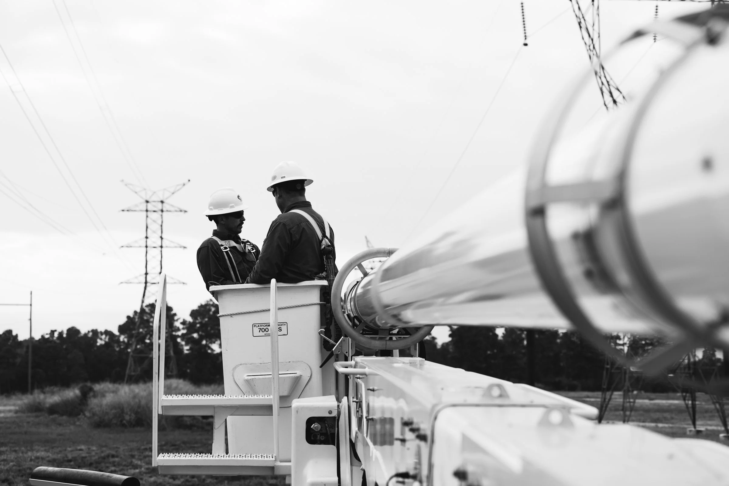 Two utility workers in safety helmets and harnesses are working on a high-voltage power line from an elevated bucket lift, with power lines and electrical transmission towers in the background.