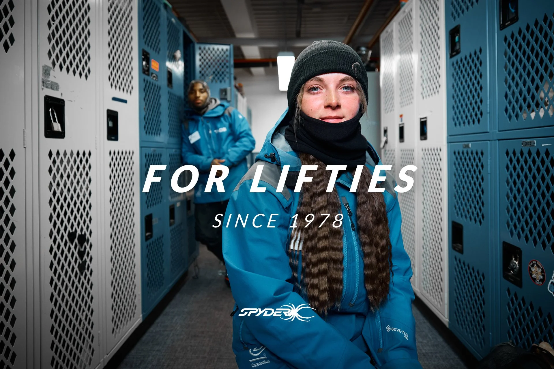Two young women in blue jackets and black beanies in a locker room, one sitting and the other standing in the background.
