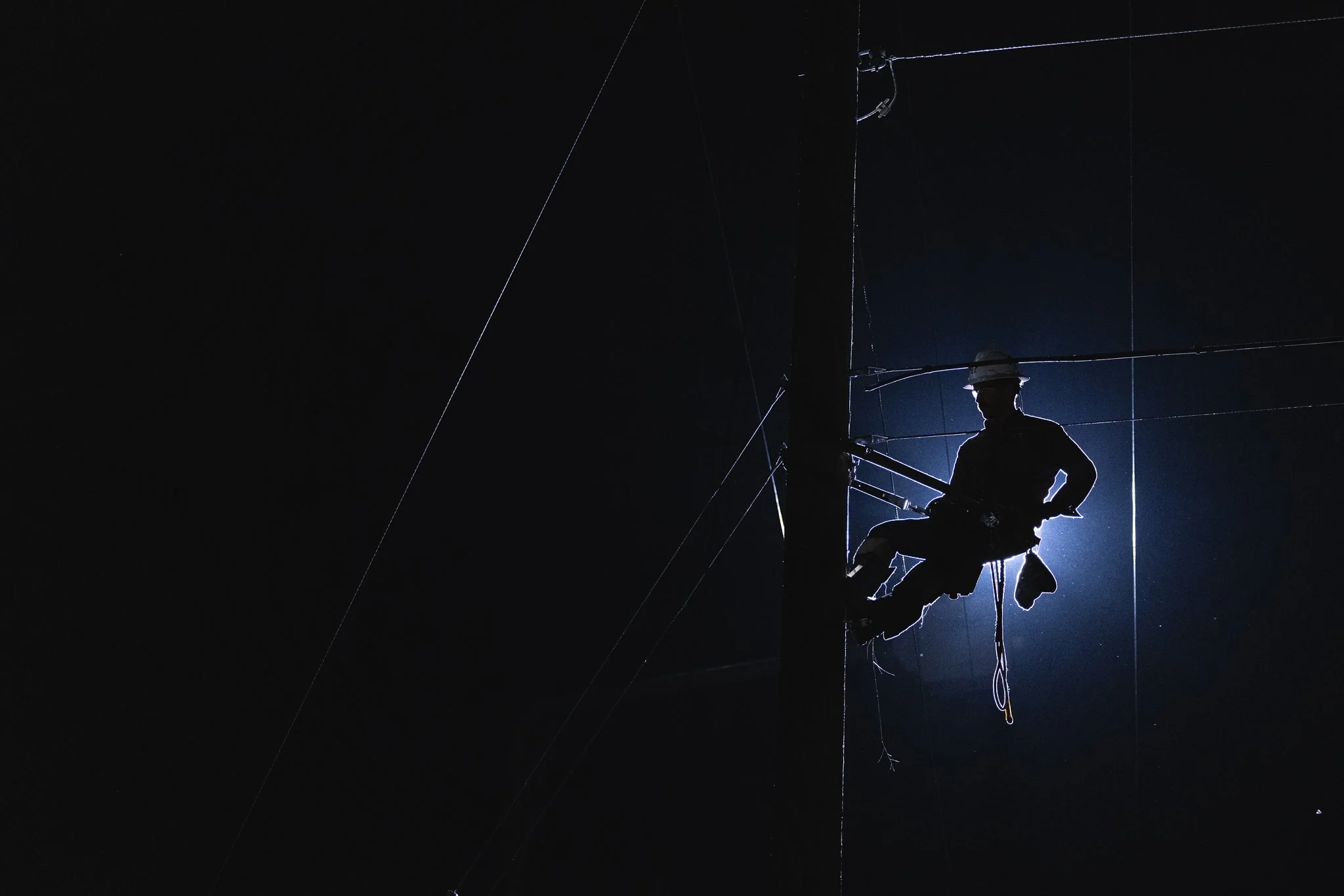 Silhouette of a lineman working at night on power lines, backlit by a bright light.