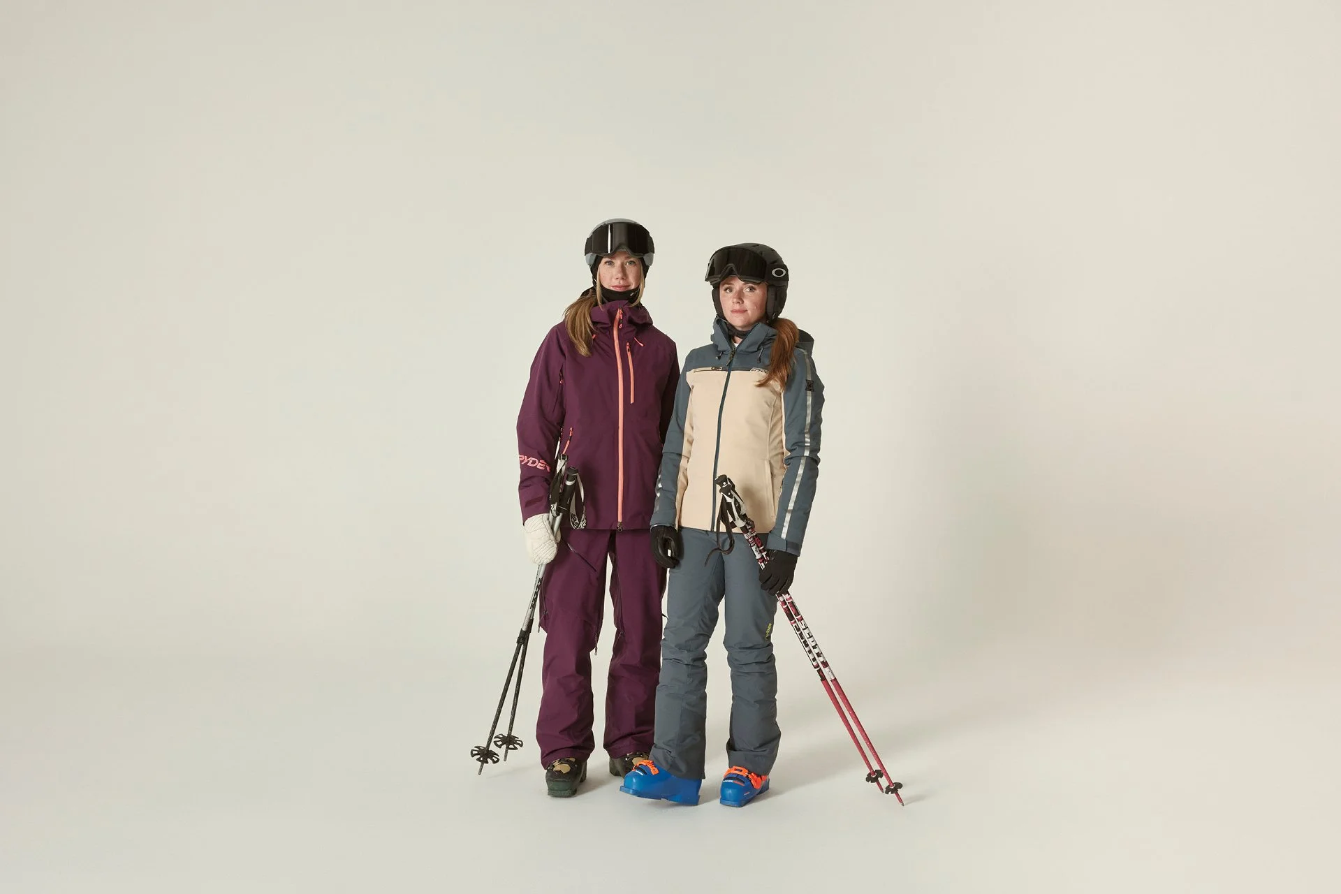 Two women dressed in ski gear, standing with ski poles against a plain white background.