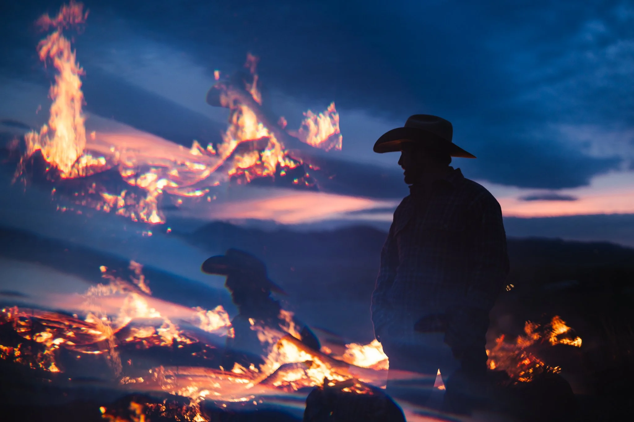 Silhouette of a person wearing a cowboy hat standing in front of a wildfire at dusk.