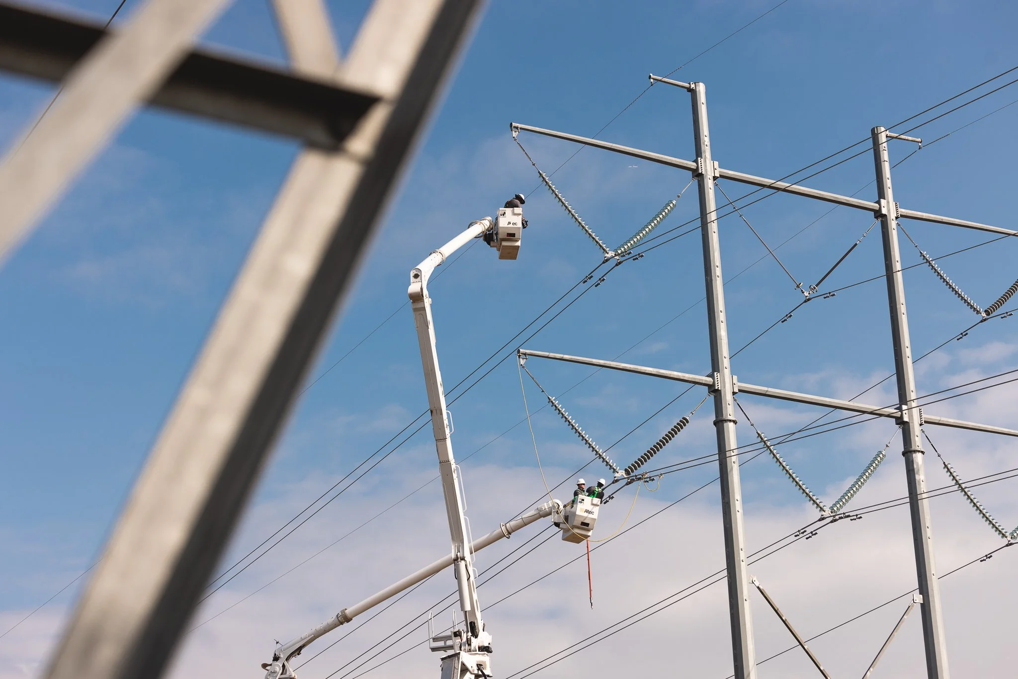 Workers in a bucket lift repairing electrical power lines on a utility pole against a blue sky.