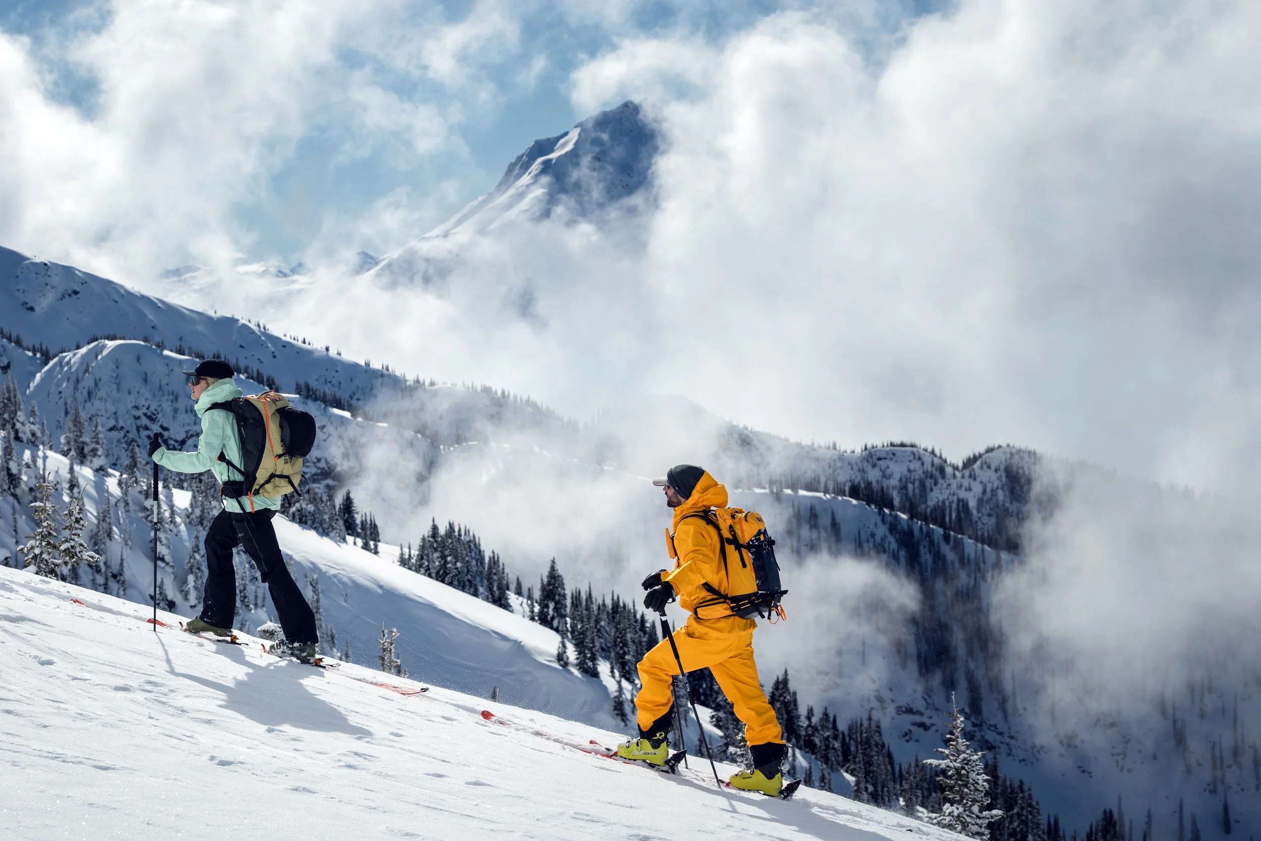 Two people skiing on a snowy mountain slope with a backdrop of snow-covered trees, mountain peaks, and clouds.