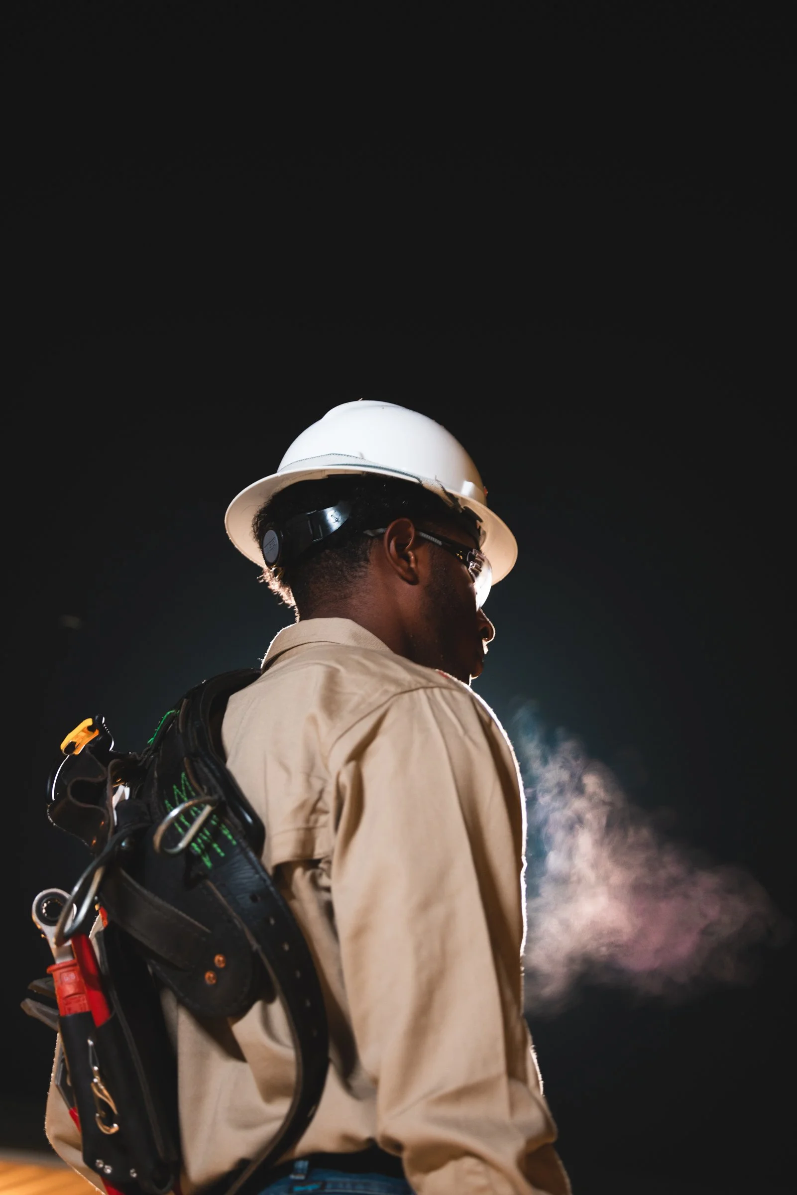 A construction worker wearing a white helmet, glasses, and beige work clothes, standing outdoors at night with visible breath in the cold.