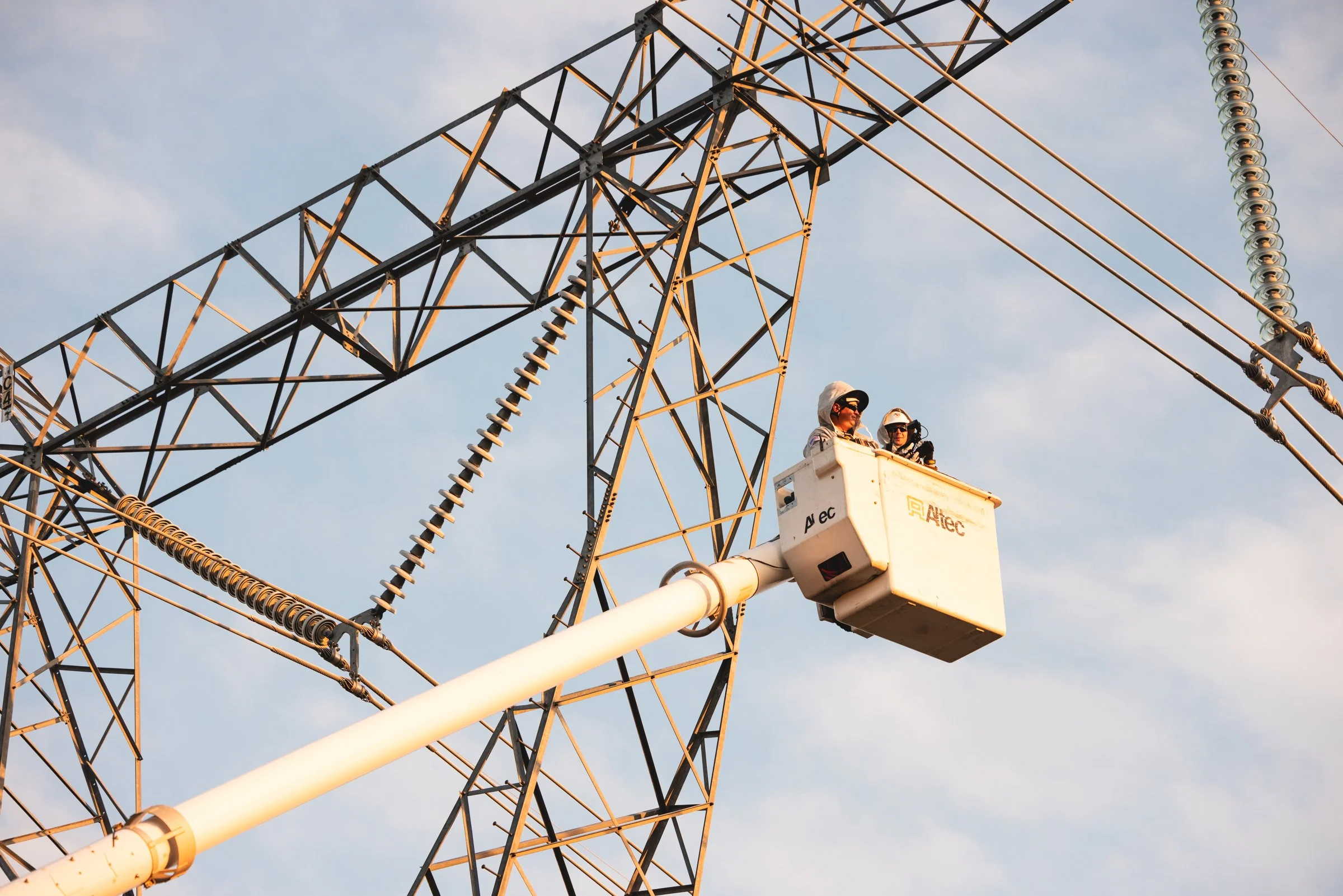 Two workers in a bucket lift inspecting high-voltage power lines on a steel electricity transmission tower during daylight.