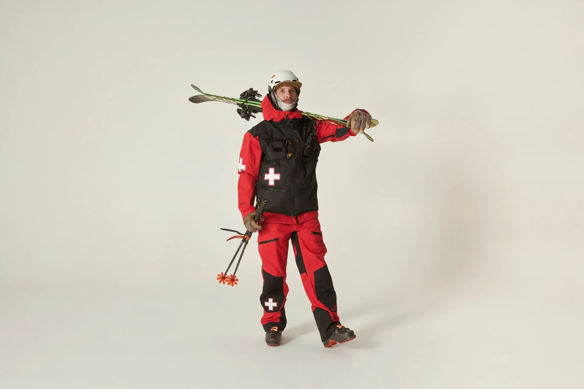 A person dressed as a ski rescue worker holding skis and ski poles, wearing red and black winter gear with a white cross on the sleeves, standing against a plain white background.
