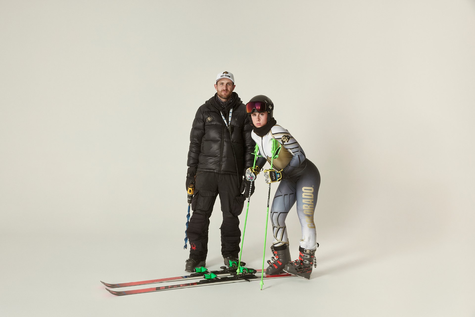A woman dressed in ski gear standing next to a man in winter clothing, both with skis and poles against a plain white background.