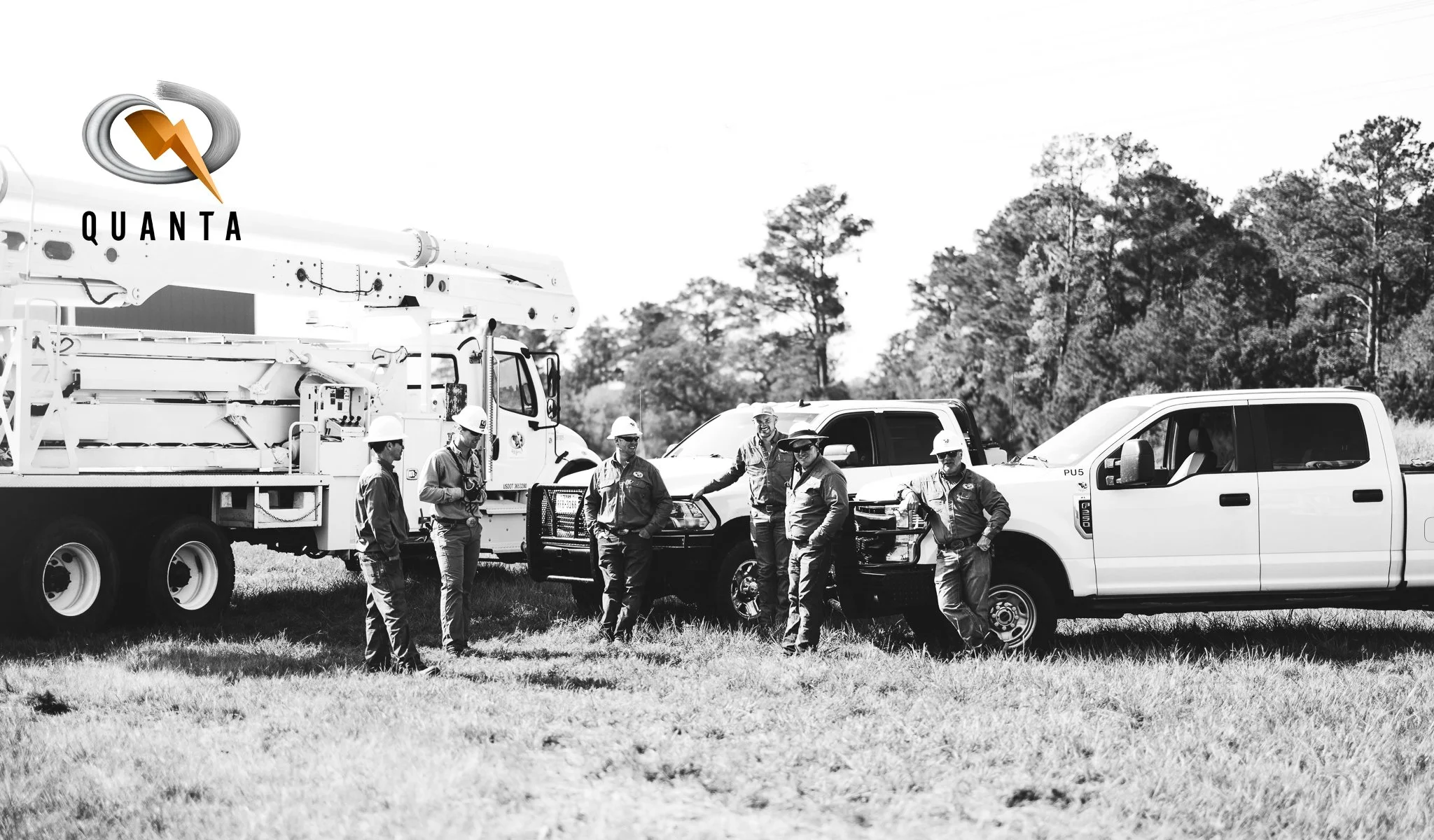 Group of workers wearing safety helmets and uniforms standing near trucks and equipment in an open field with trees in the background.