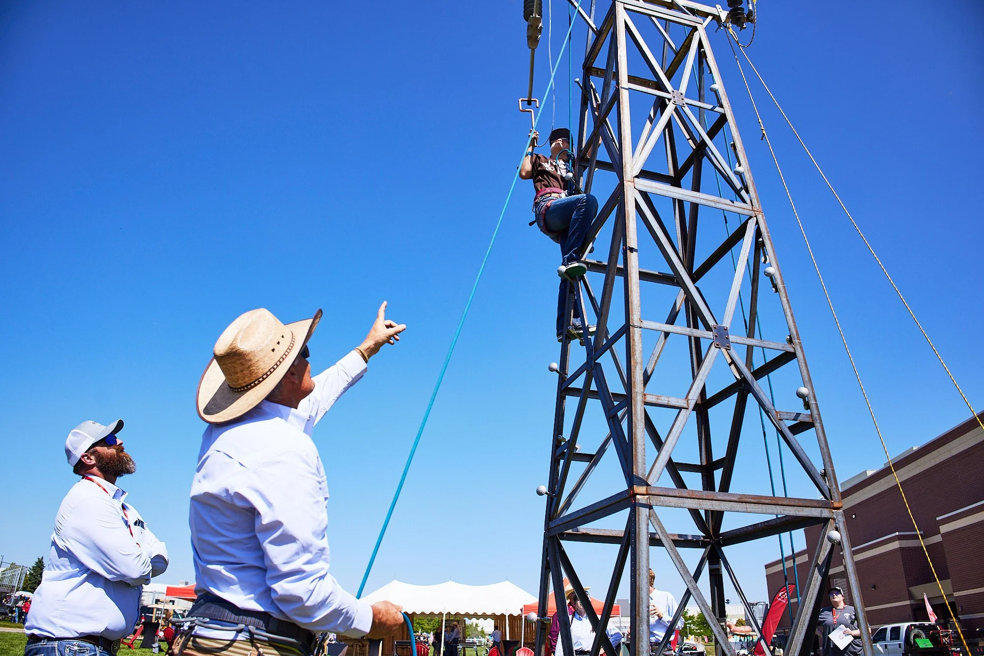 Child wearing a harness climbing a metal tower at an outdoor event, with two adults observing and one adult pointing, under a clear blue sky.