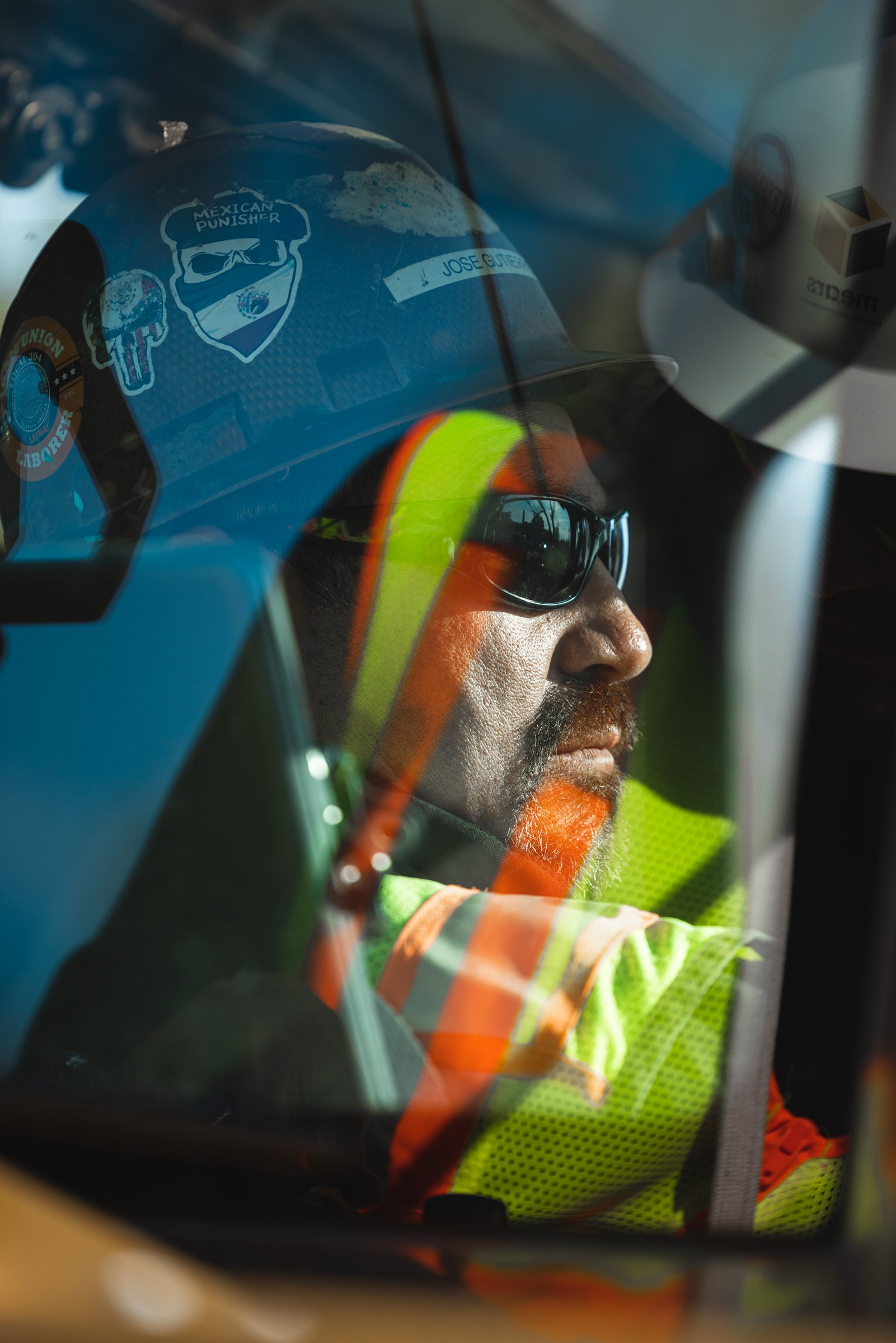A man wearing sunglasses, a high-visibility safety vest, and a helmet with stickers, seen through a vehicle windshield.