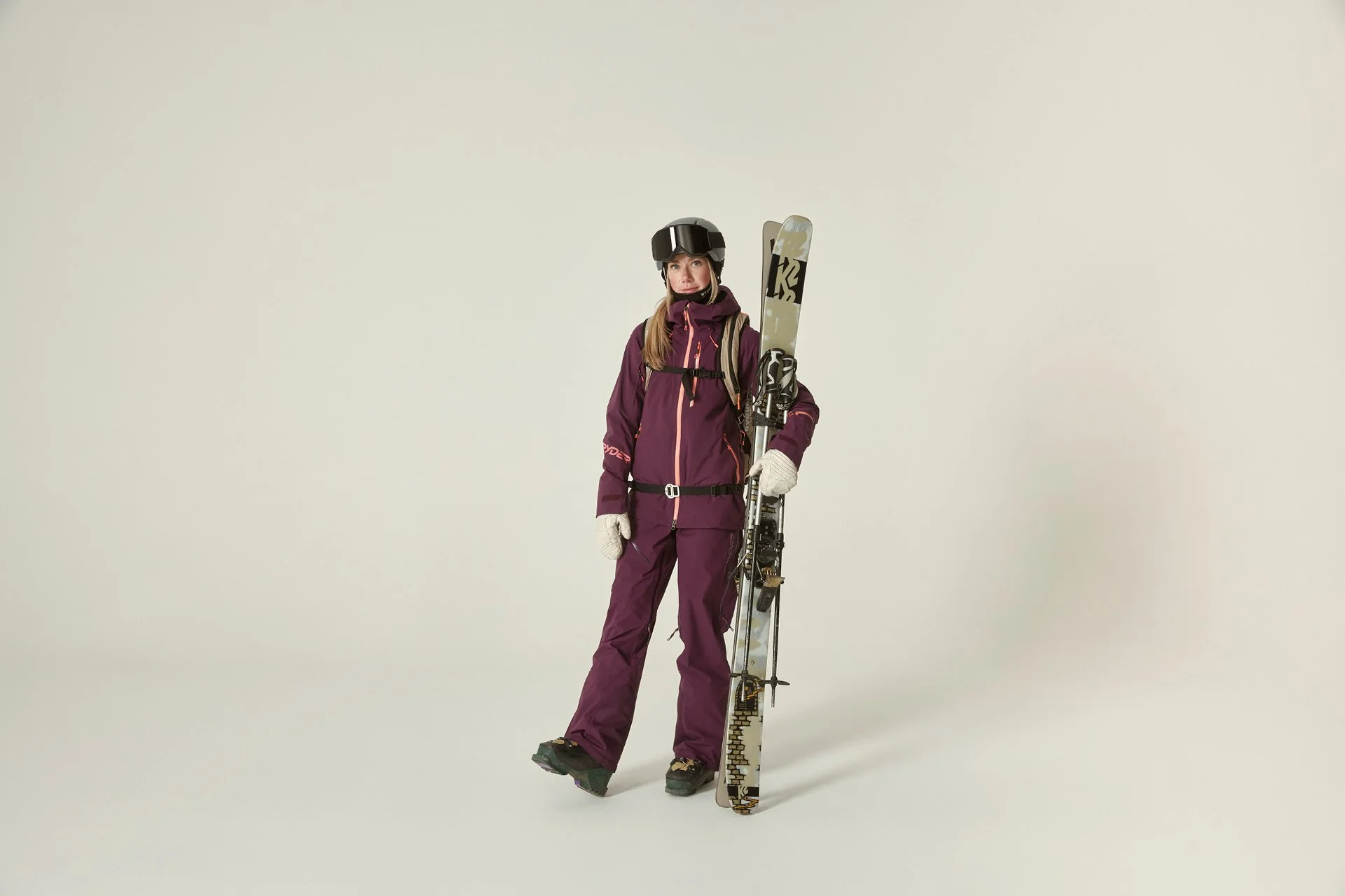 A woman dressed in purple ski gear holding skis and ski poles, wearing a helmet and gloves, standing against a plain light background.